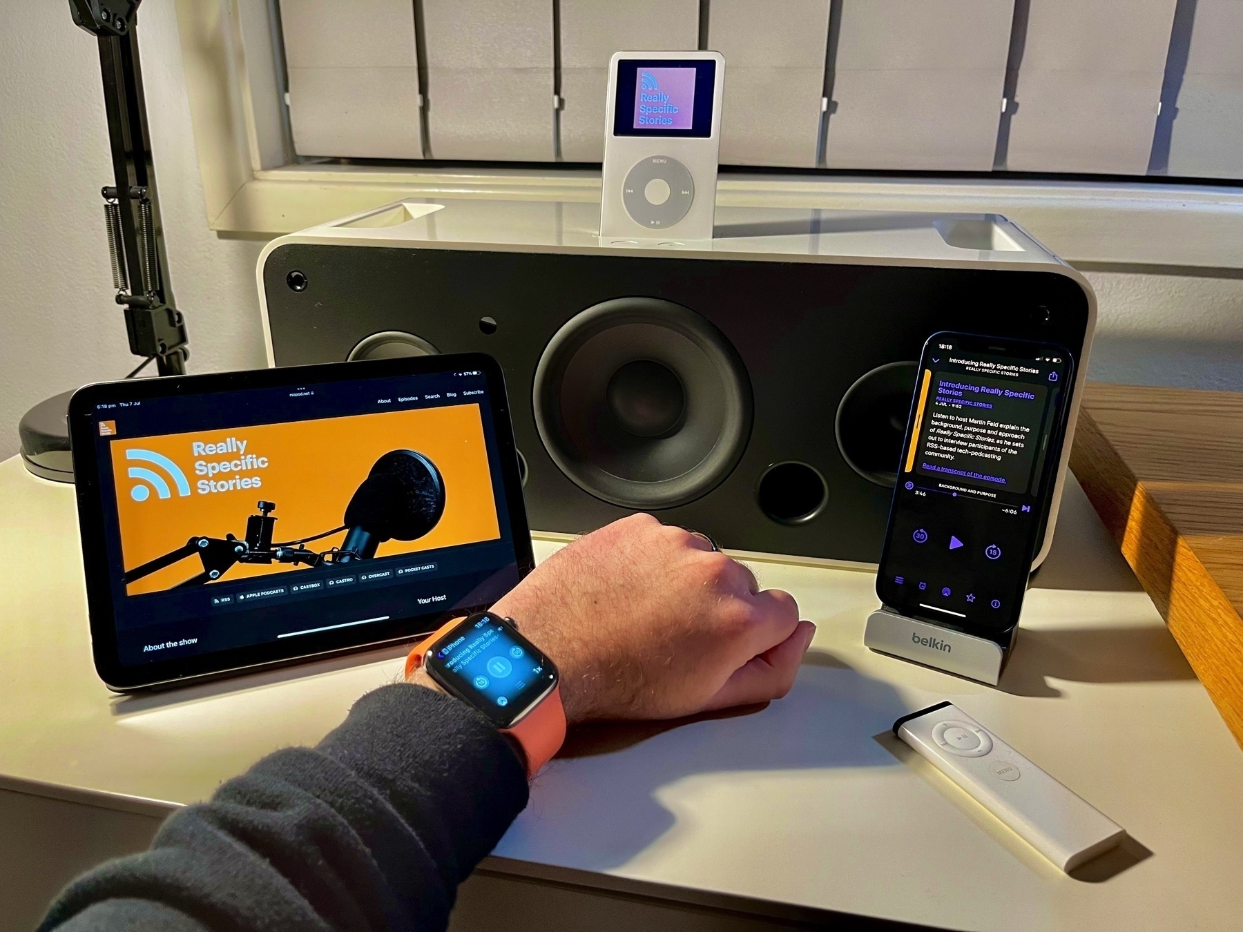 Podcast artwork and apps displayed across an Apple Watch, iPad mini (6th gen.), iPhone 12 mini and white iPod Video docked in an iPod Hi-Fi