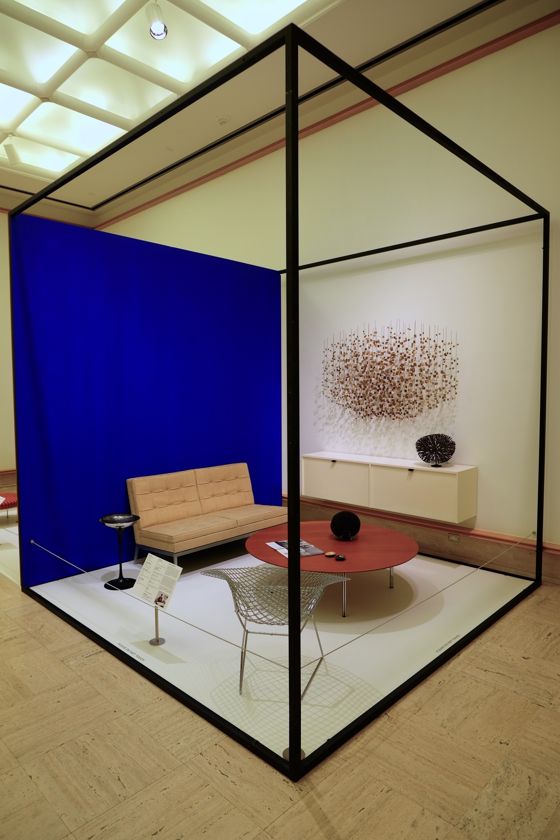 A modern furniture display features a minimalist seating area with a red circular table, a beige couch, a wire chair, and a striking wall sculpture, all enclosed within a black metal frame structure.