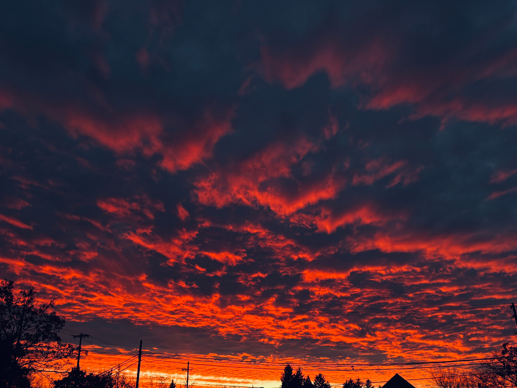 A vibrant sunset with fiery orange and red clouds dominates the sky above silhouetted trees and power lines.