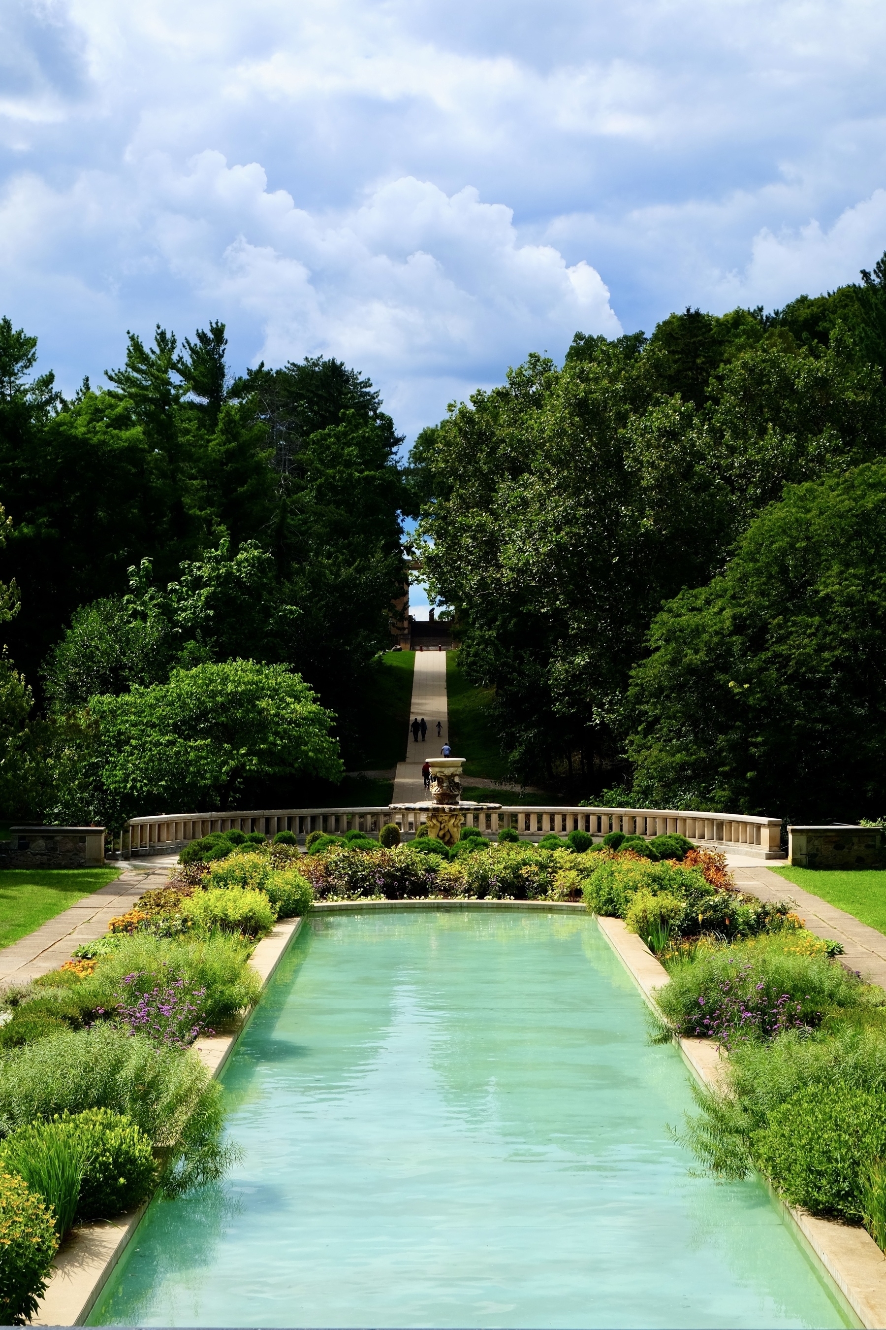 A long, rectangular reflecting pool is surrounded by lush greenery and landscaped gardens under a cloudy sky.
