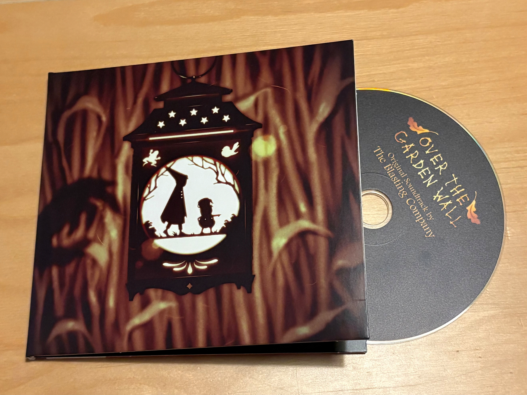A CD titled Over the Garden Wall, original soundtrack, is partially inserted in a case featuring an illustration of two silhouetted figures inside a lantern against a forest background. 