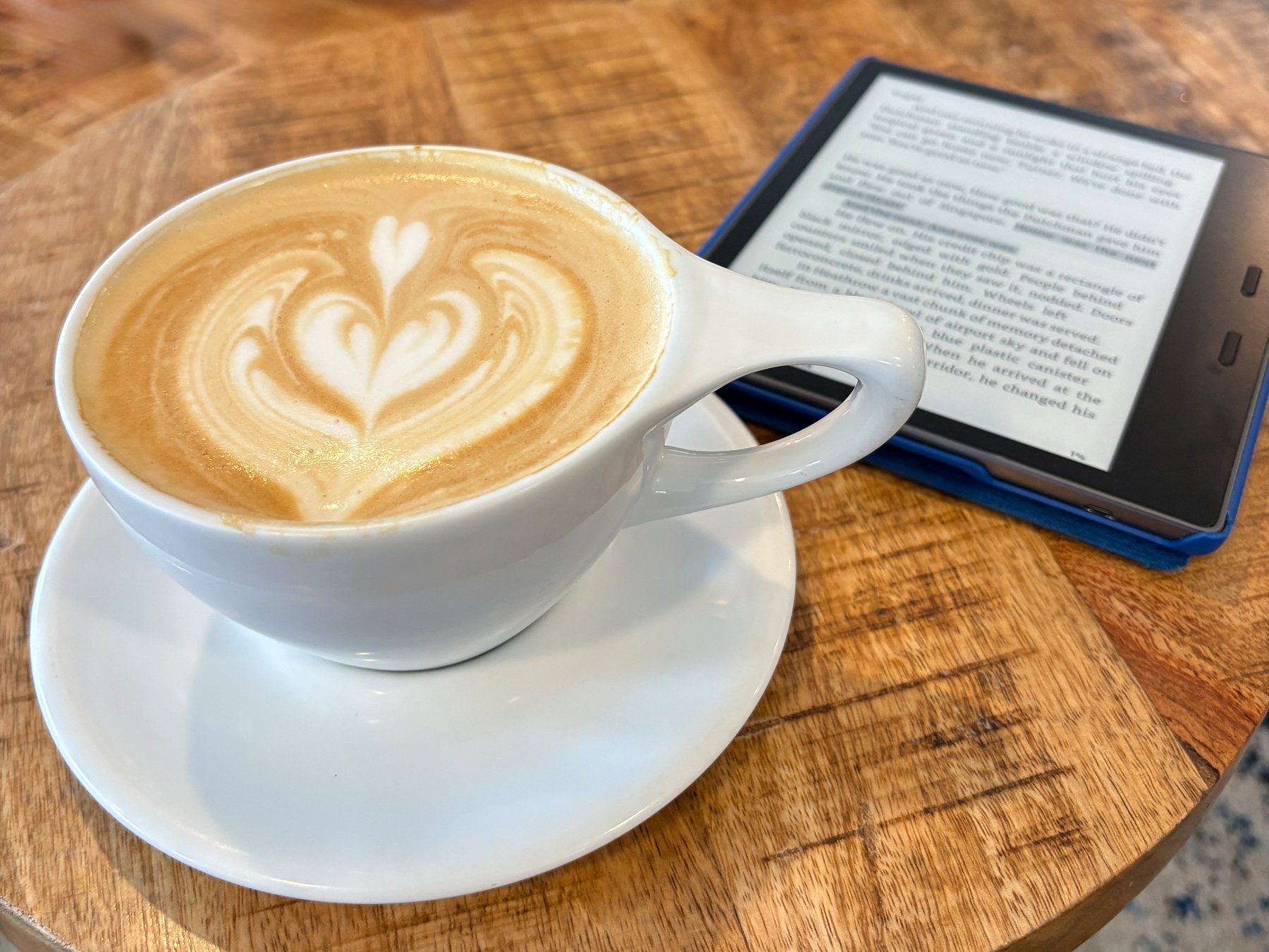 A cappuccino with heart-shaped latte art is in a white cup on a saucer next to an e-reader displaying text, all set on a wooden table.