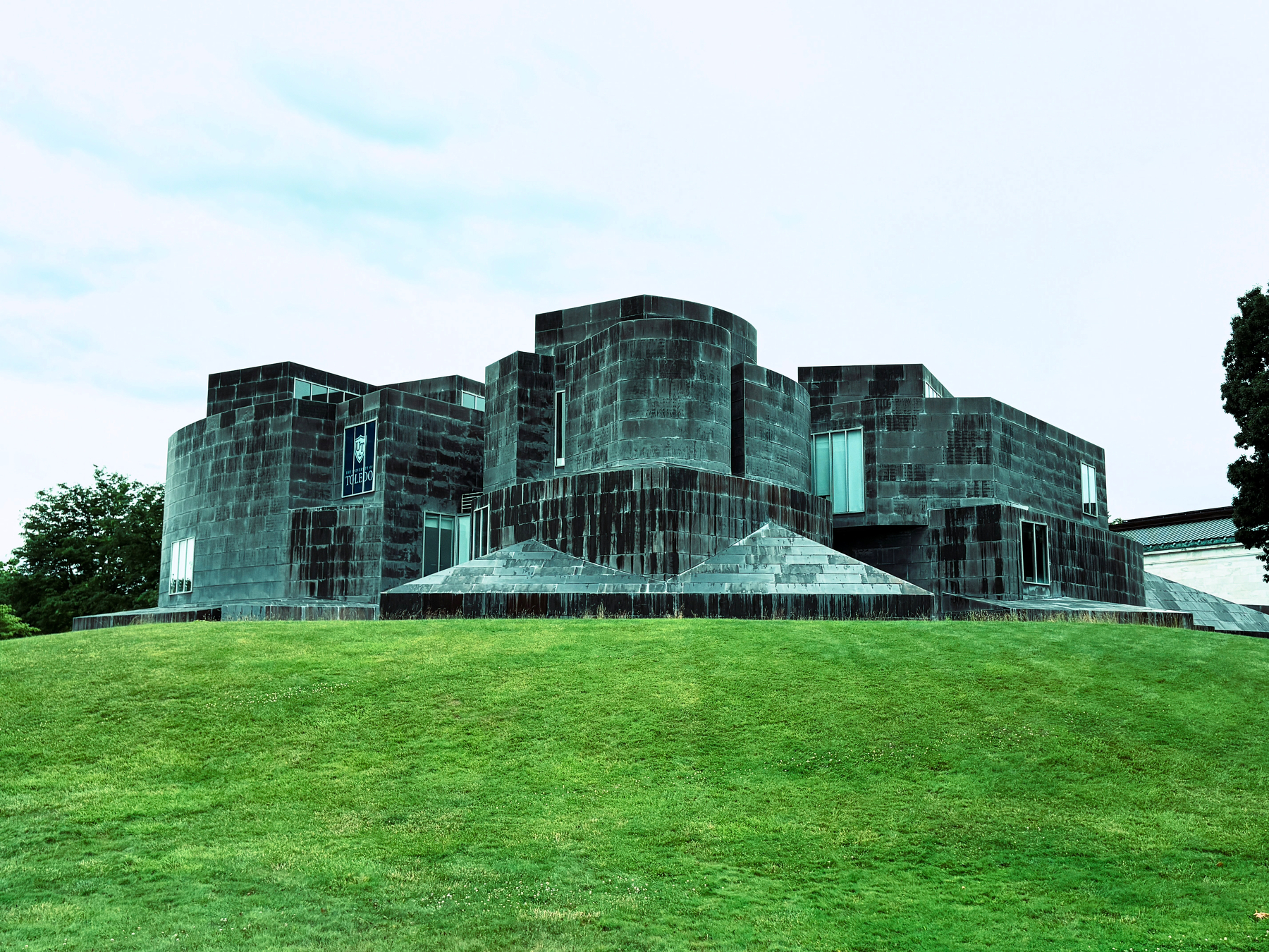 University of Toledo Center for Visual Arts. A large, modern building with curved and angular dark stone walls is situated on a grassy hill under a cloudy sky.