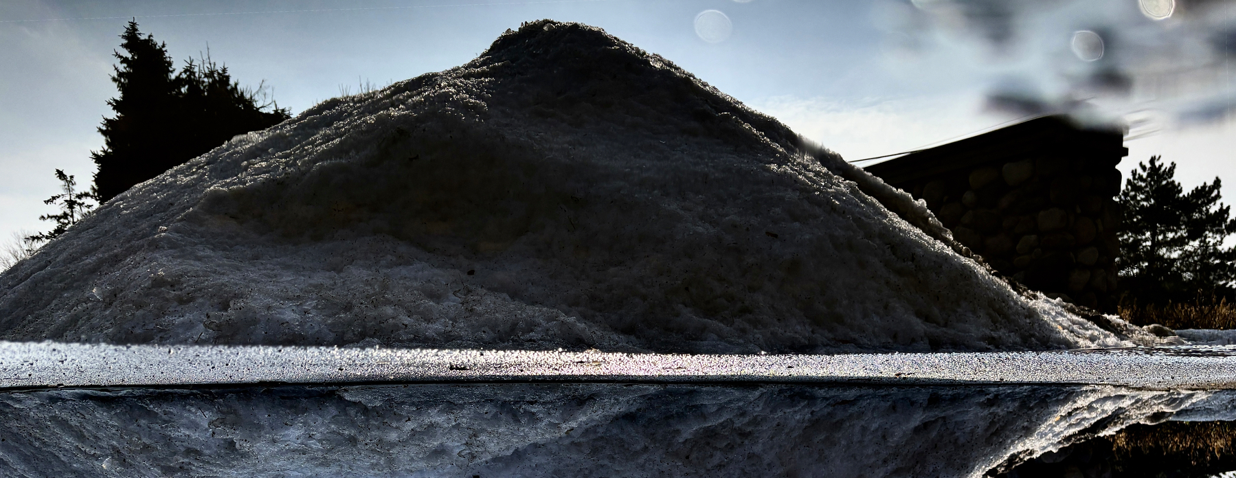 A large mound of snow casts a shadow on the ground, reflecting in a nearby puddle under a partly cloudy sky.