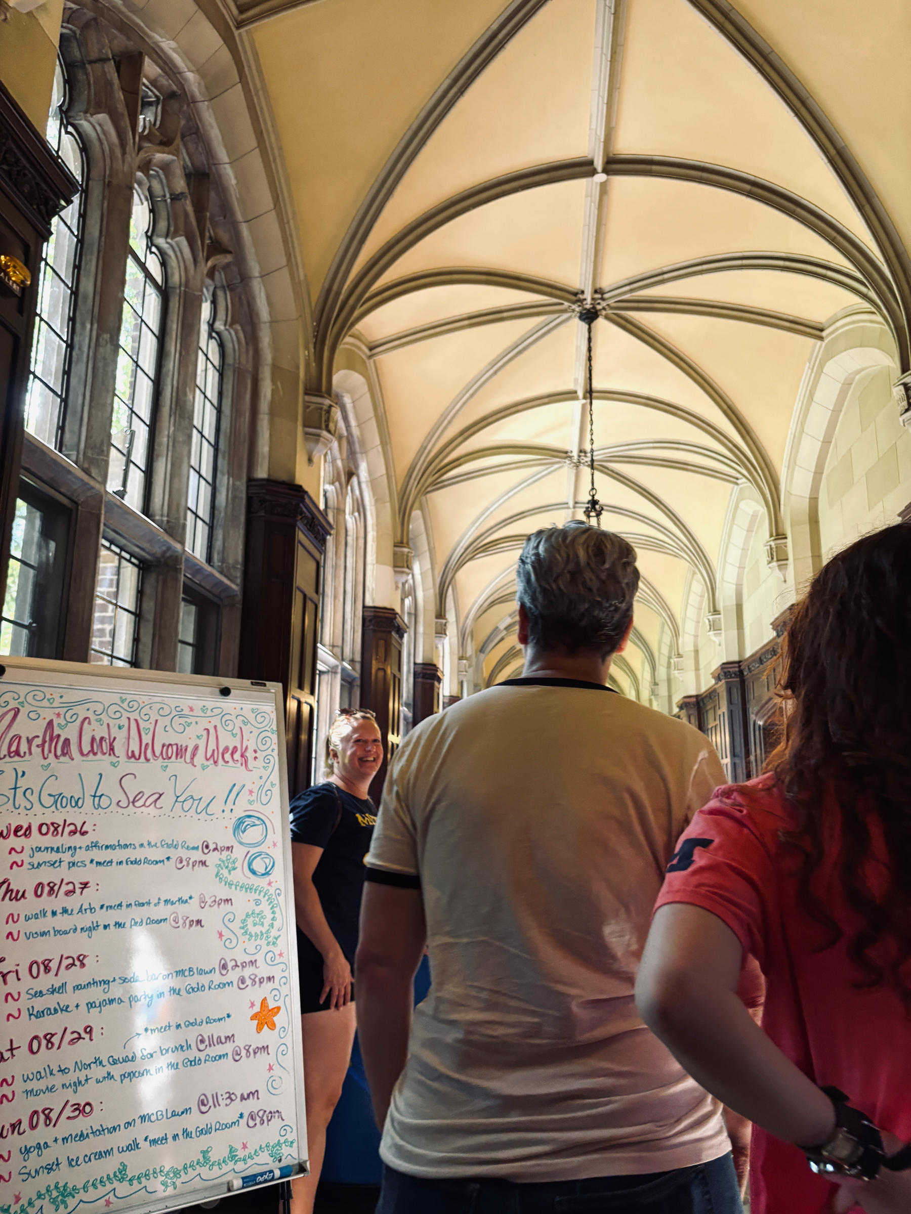 People are gathered in a hallway with arched ceilings, near a colorful signboard detailing a college welcome week schedule.