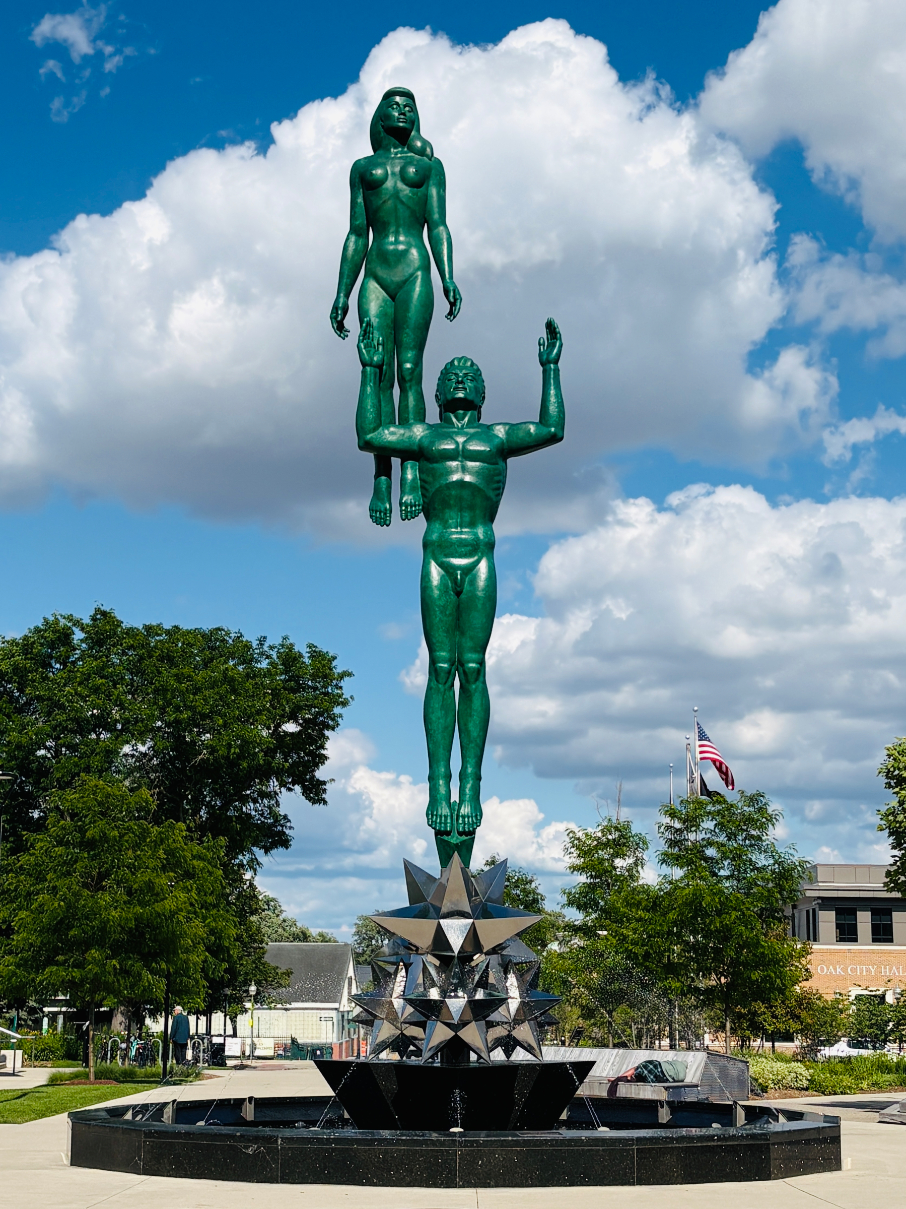 A green statue of a man holding a woman above his head stands on a geometric pedestal in an outdoor park in Royal Oak, Michigan with trees and buildings in the background.