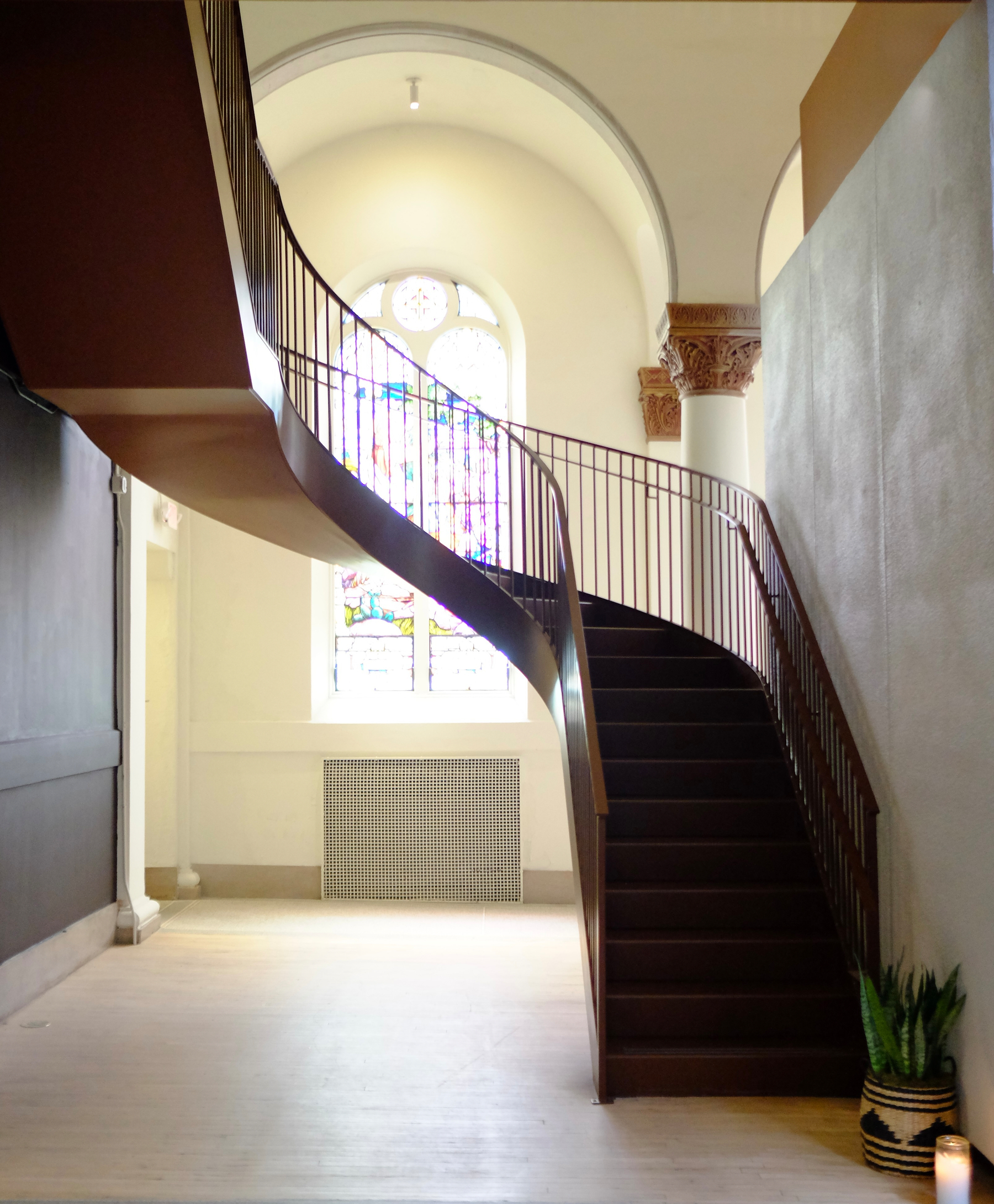 A curved staircase with a metal railing is positioned in a bright, airy interior featuring arched stained glass windows and ornate architectural details.
