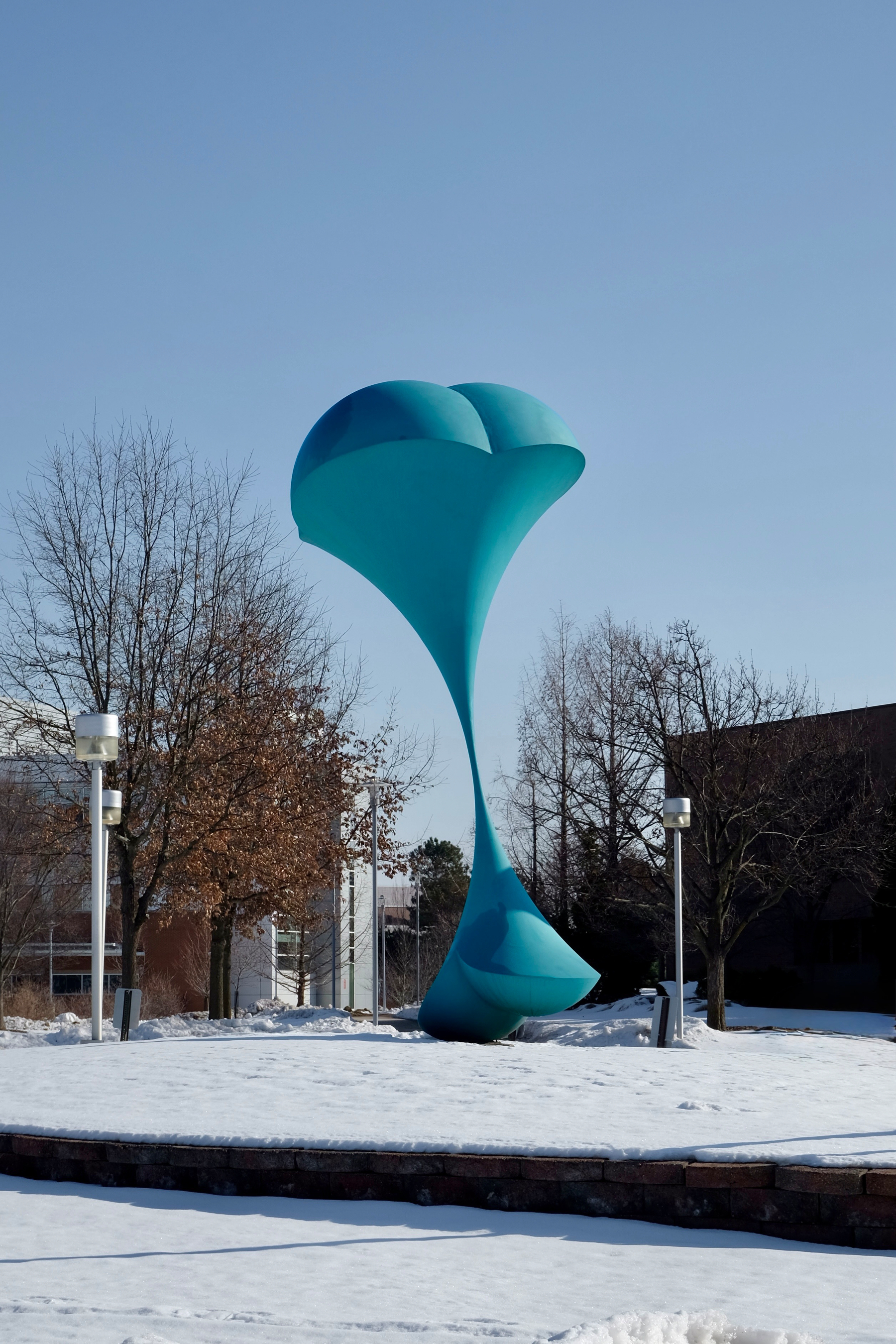 A large blue abstract sculpture stands on a snowy outdoor plaza surrounded by bare trees.