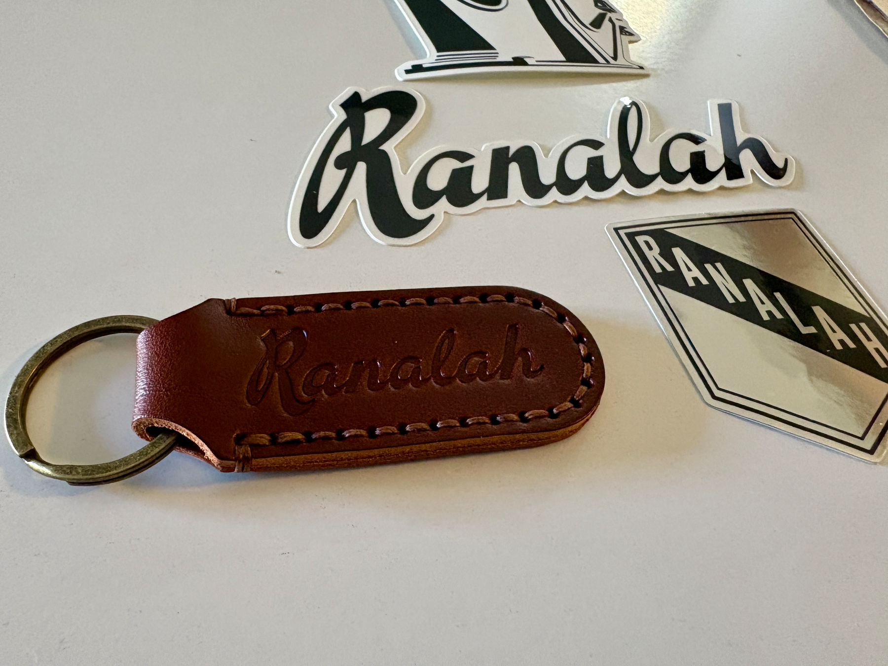 A brown leather keychain and several stickers, all featuring the word Ranalah, are displayed on a white surface.