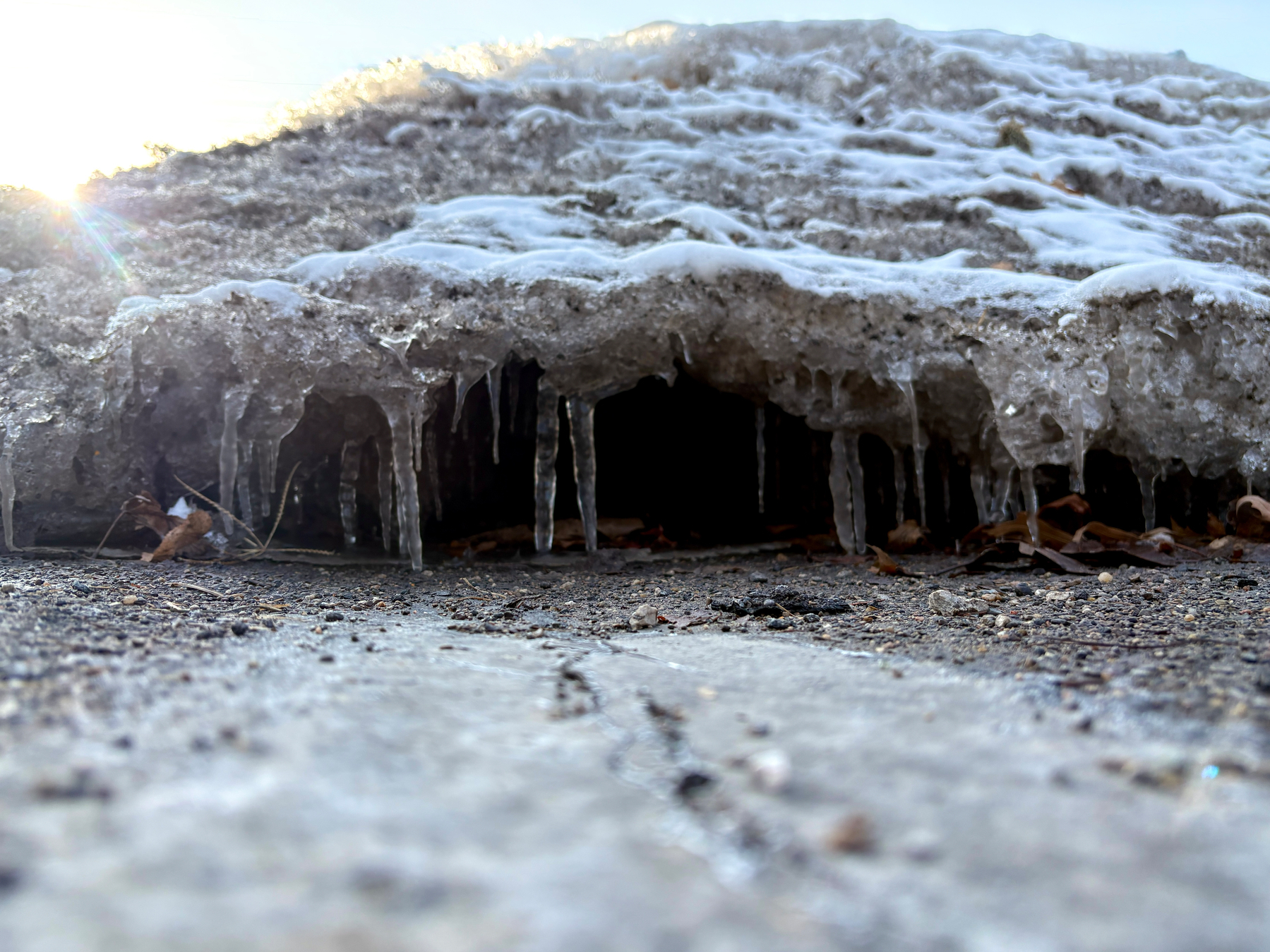 A layer of ice resembling a cave formation, with icicles hanging from it, sits on a rough ground surface.