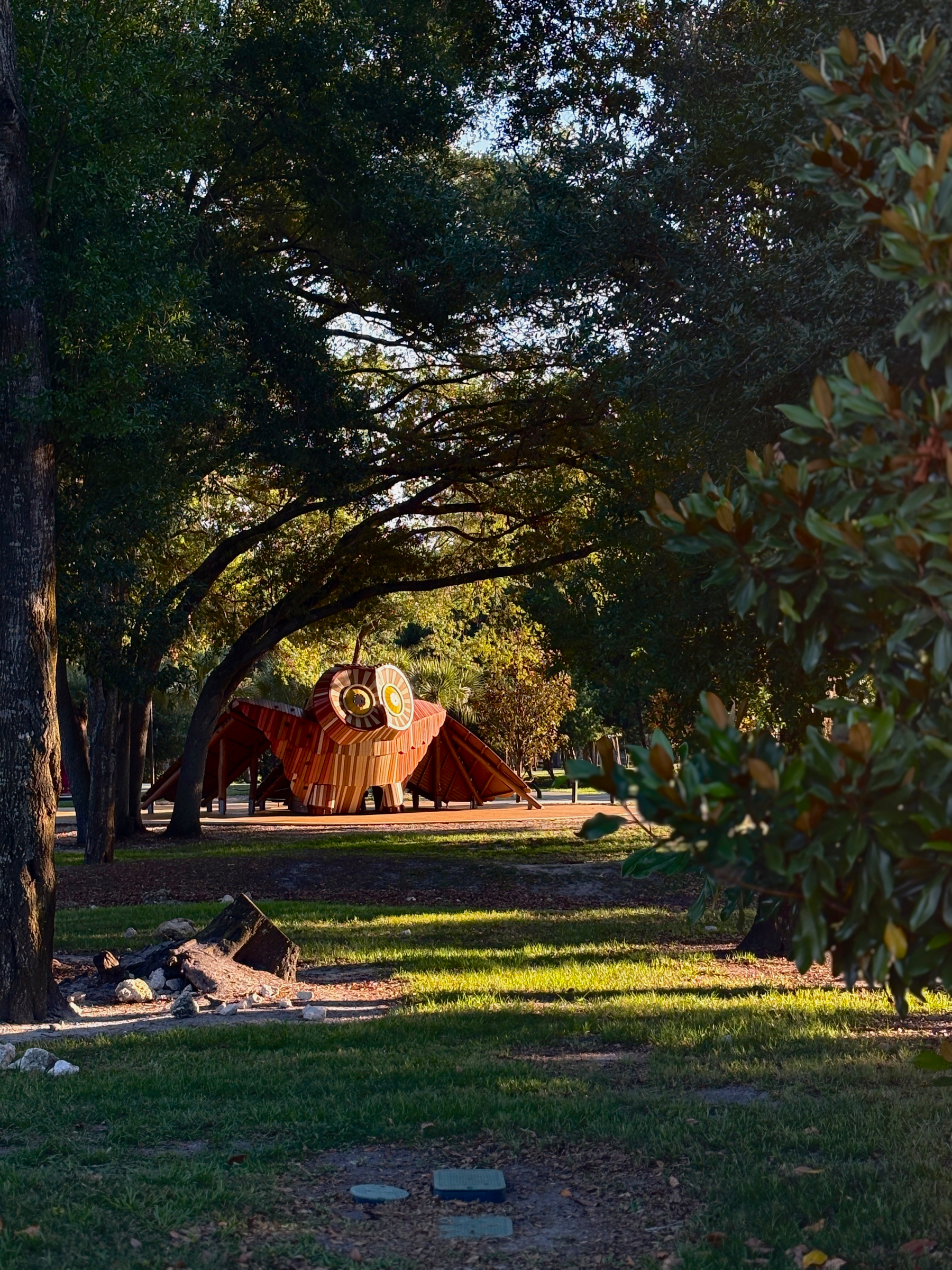 A large wooden owl sculpture is nestled among trees in a sunlit Bonnet Springs park setting.