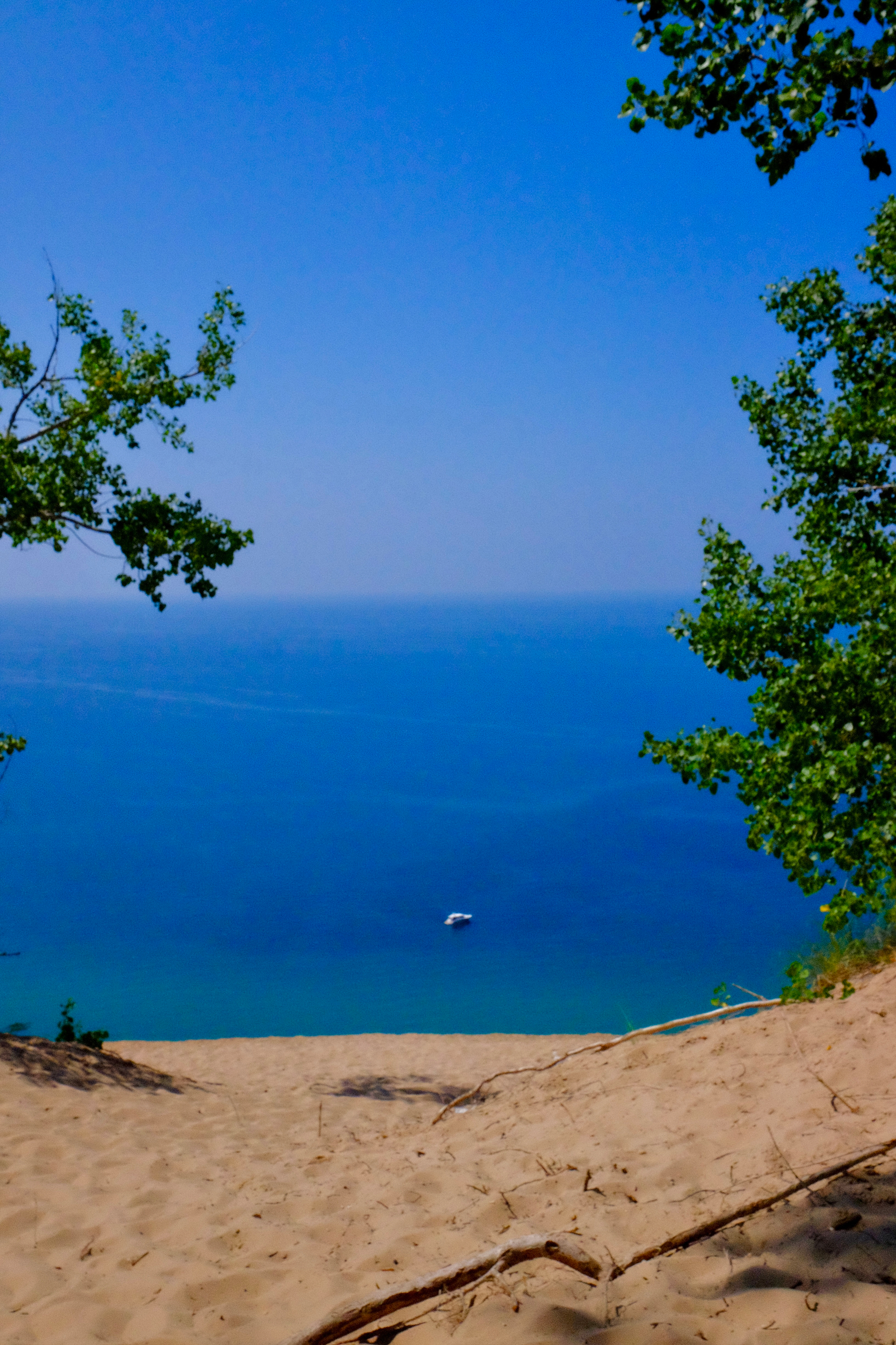 A sandy path at Sleeping Bear Dunes bordered by green trees leads down to a vast, calm body of blue water with a small boat floating in the distance.