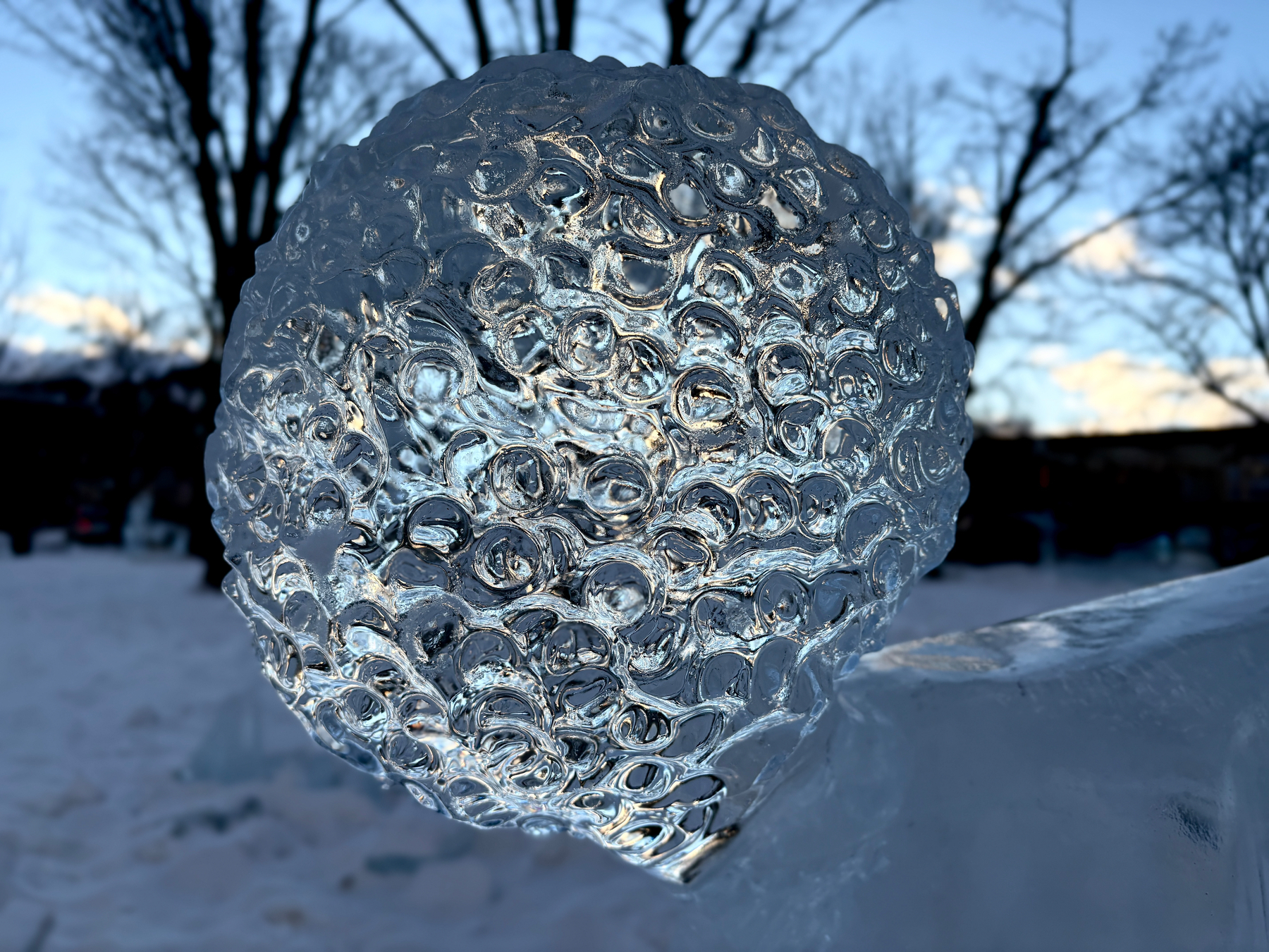 A textured ice sculpture shaped like a sphere is set against a snowy and tree-lined background.