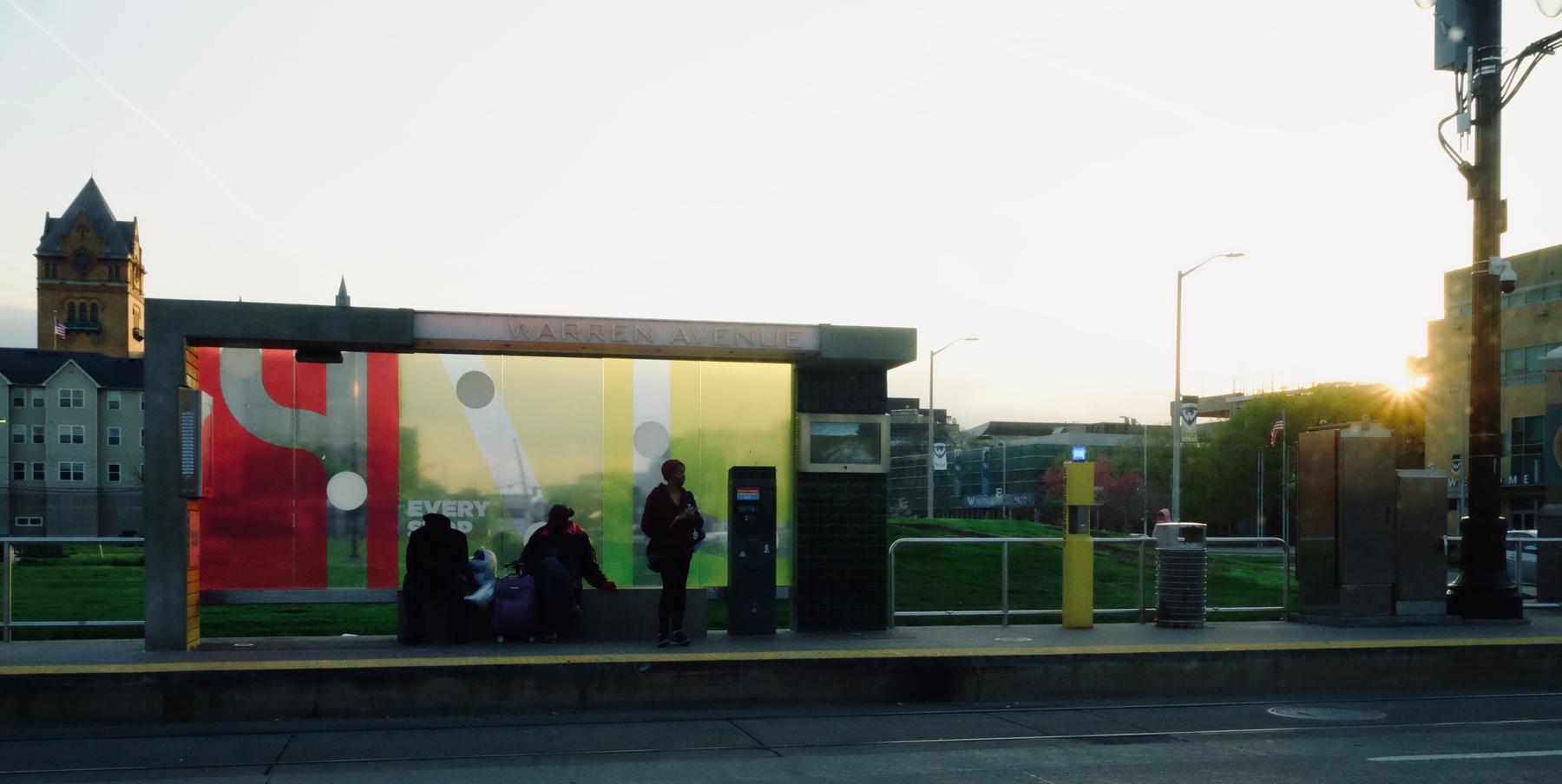 Several people are sitting or standing at a colorful bus shelter during sunset, with buildings visible in the background.