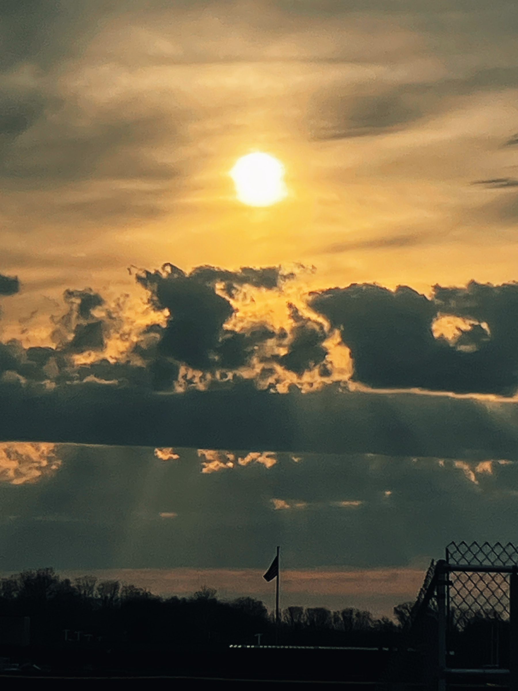 A bright sun sets behind dramatic clouds above a silhouetted landscape with a flag and fence.