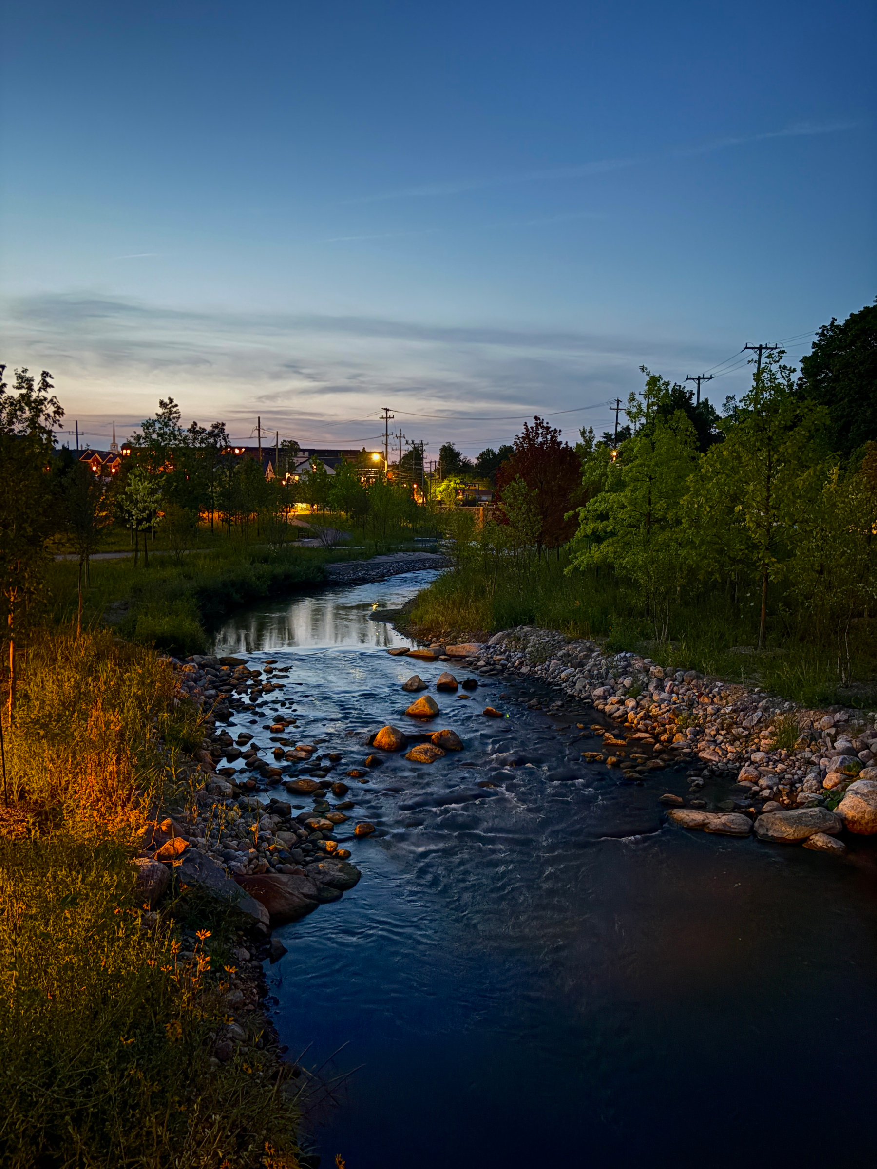A gently flowing river bordered by rocks and lush greenery winds through a quiet area with houses and power lines visible under a twilight sky.