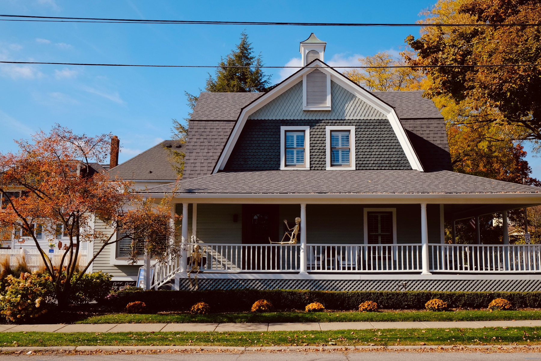 A charming two-story house with a large front porch is surrounded by autumn trees and shrubbery. Two skeletons sit on the porch, waving.