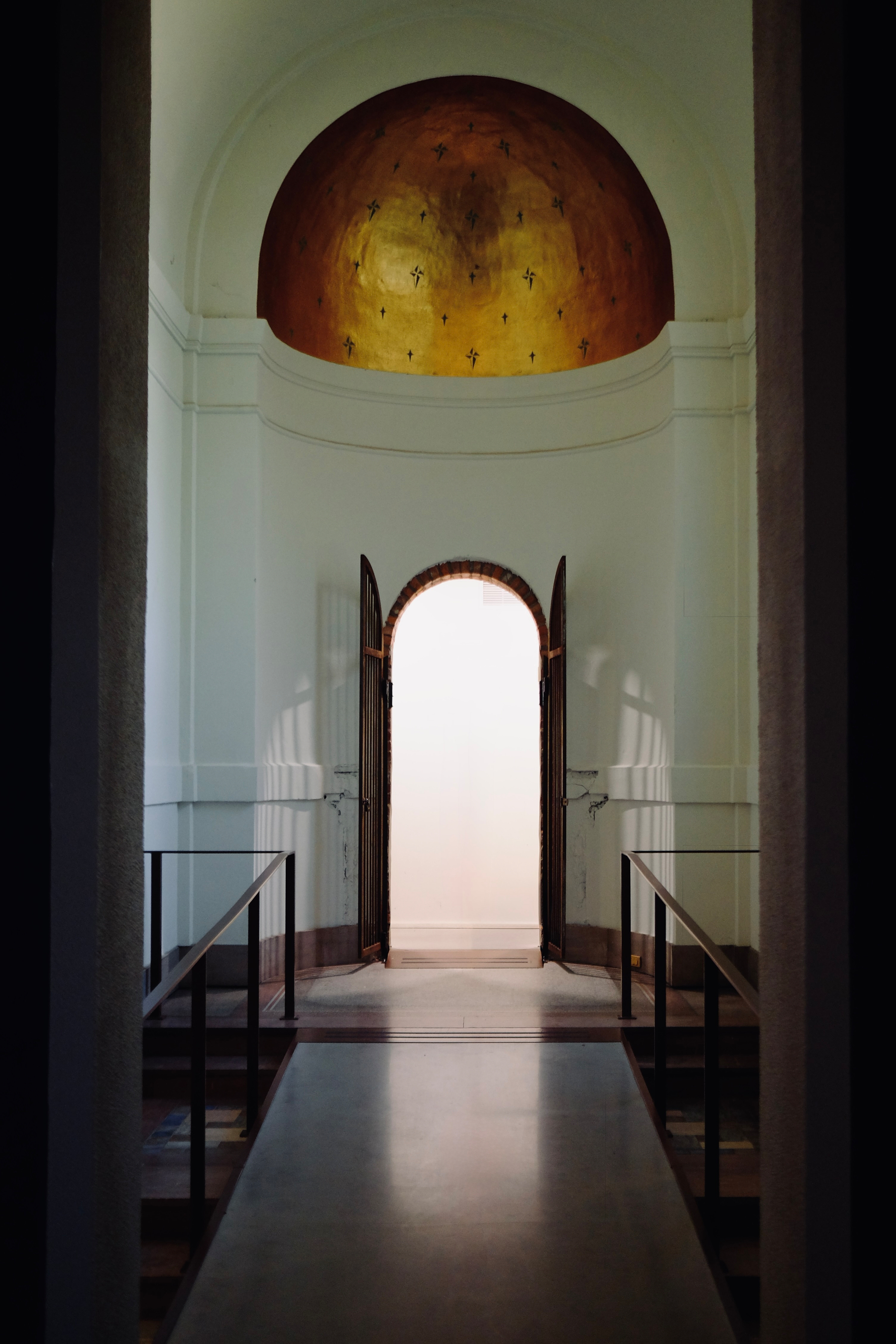 A hallway leads to a wooden double door with an arched top, set beneath a golden dome adorned with small crosses.