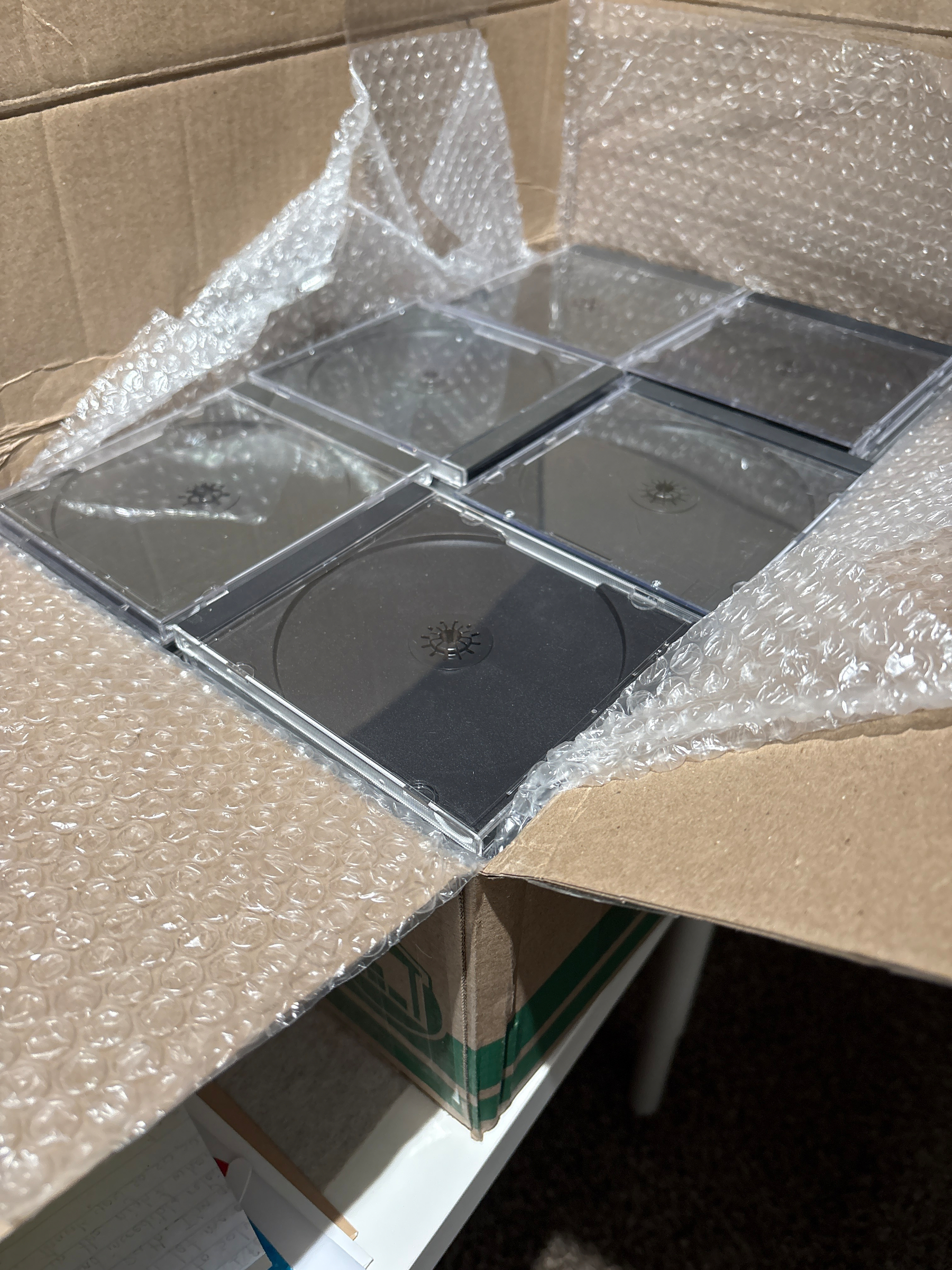 A cardboard box contains empty black CD jewel cases cushioned by bubble wrap.