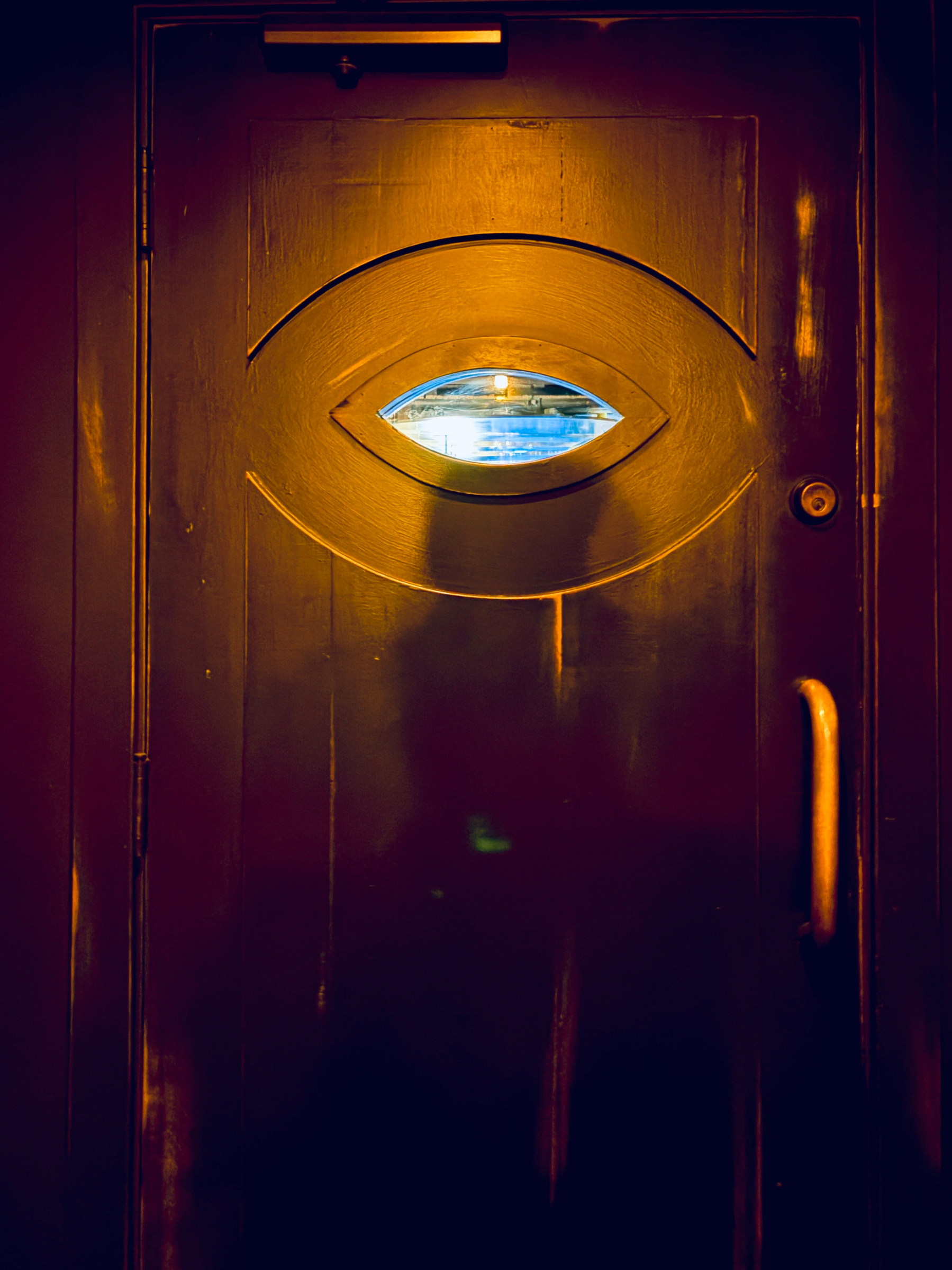 A dark wooden door features a small oval window in the center resembling an eye, with warm lighting casting shadows around it.