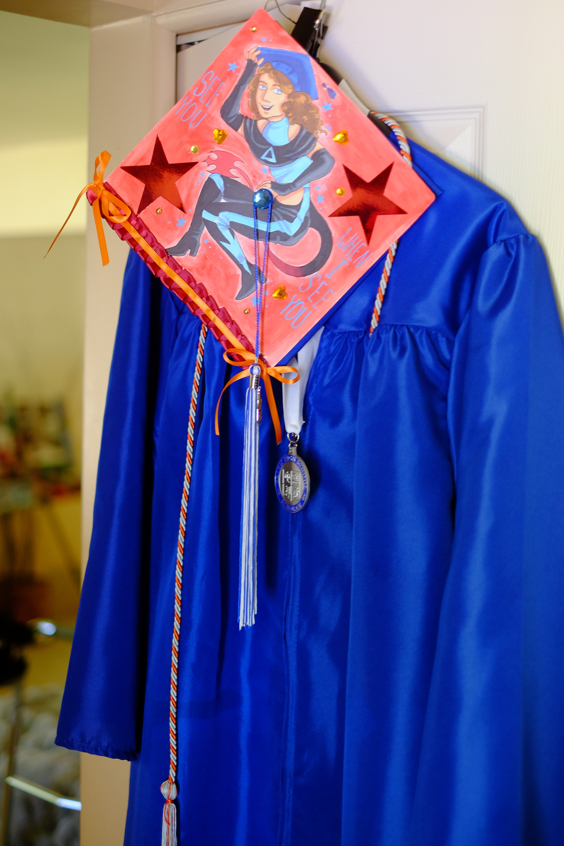 A blue graduation gown is hanging on a door, featuring a decorated cap with a painted design and colorful ribbons.