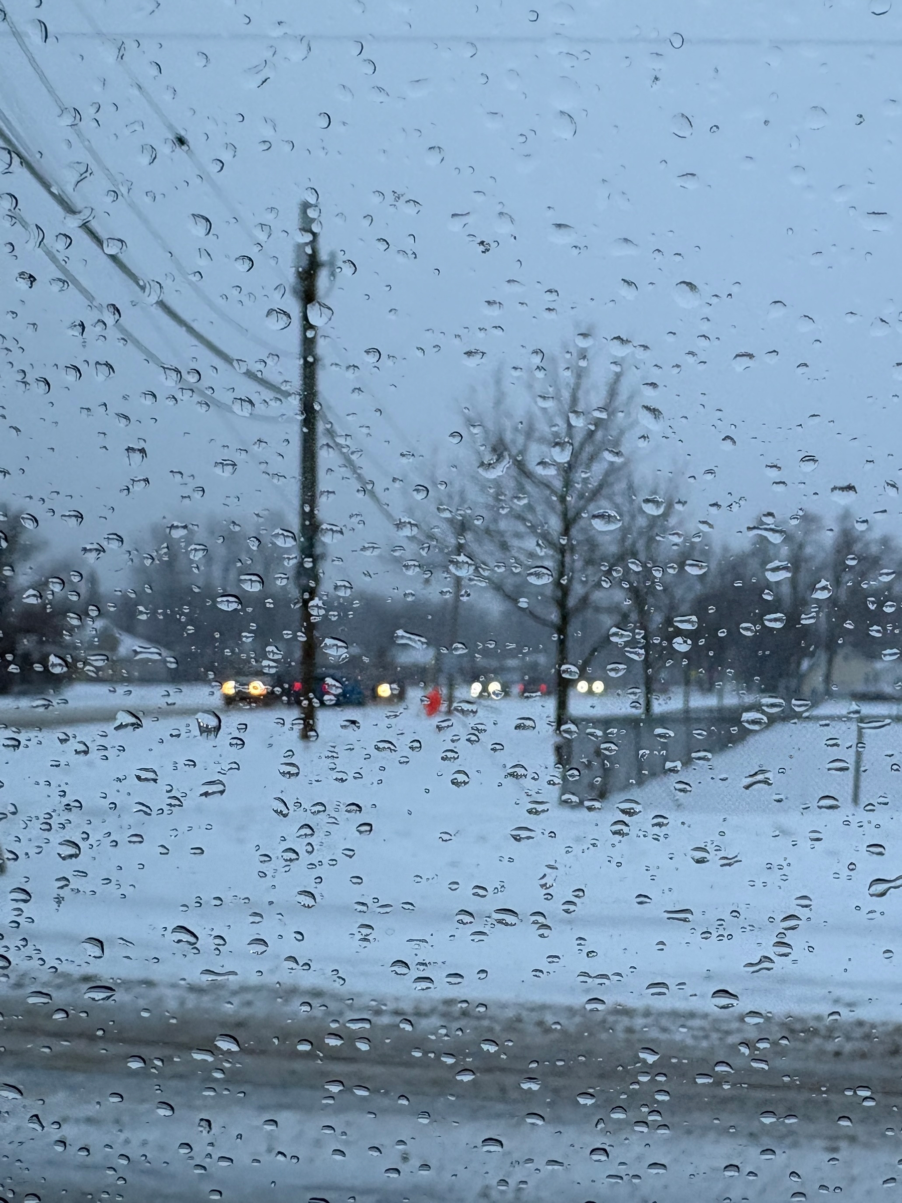 Raindrops on a window blur the view of a snowy street with trees and distant cars.