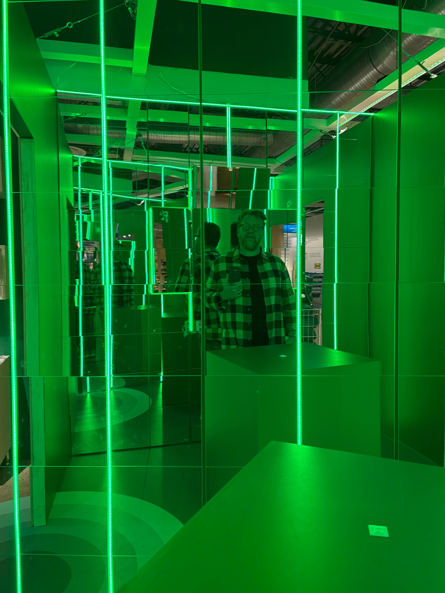 A person wearing a checkered shirt is taking a photo in a room filled with vertical green neon lights and multiple mirrors that create infinite reflections.