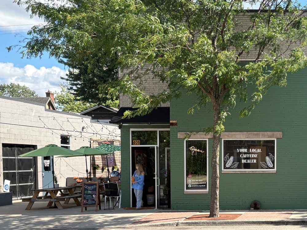 A person stands at the entrance of a green building with a sign that reads YOUR LOCAL CAFFEINE DEALER, next to outdoor seating with picnic tables and umbrellas.