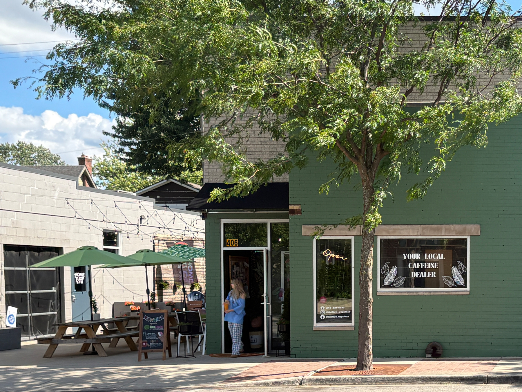 A person stands at the entrance of a green building with a sign that reads YOUR LOCAL CAFFEINE DEALER, next to outdoor seating with picnic tables and umbrellas.