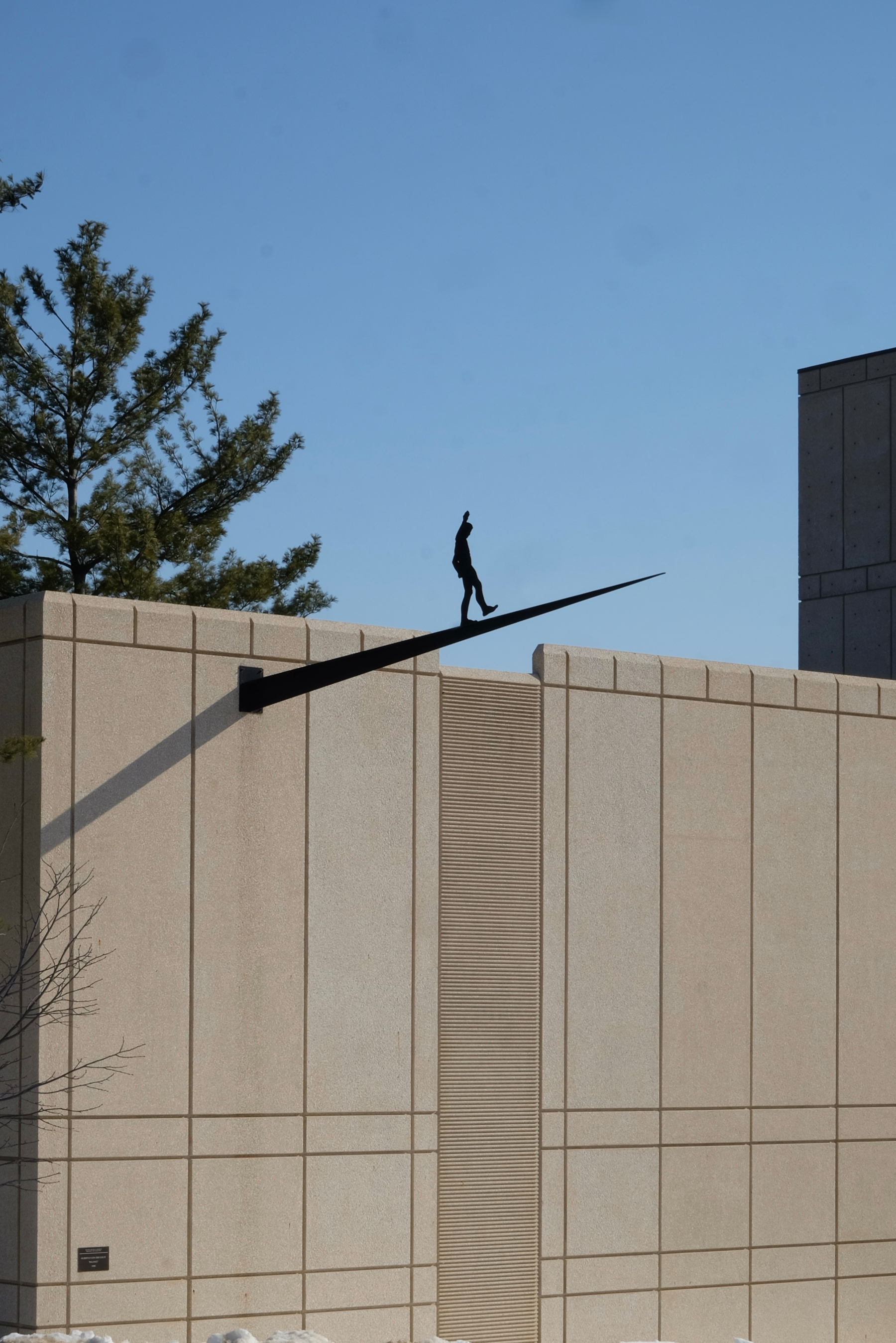 A sculpture of a person walking on a long beam protrudes from the edge of a building under a clear blue sky.