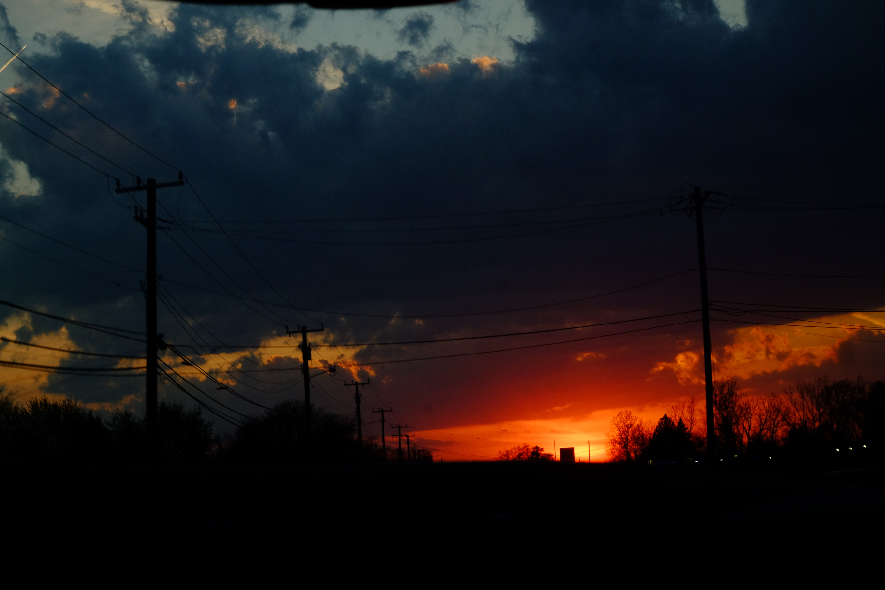 A vibrant sunset illuminates the sky with deep orange and red hues, silhouetting power lines and trees in the foreground.