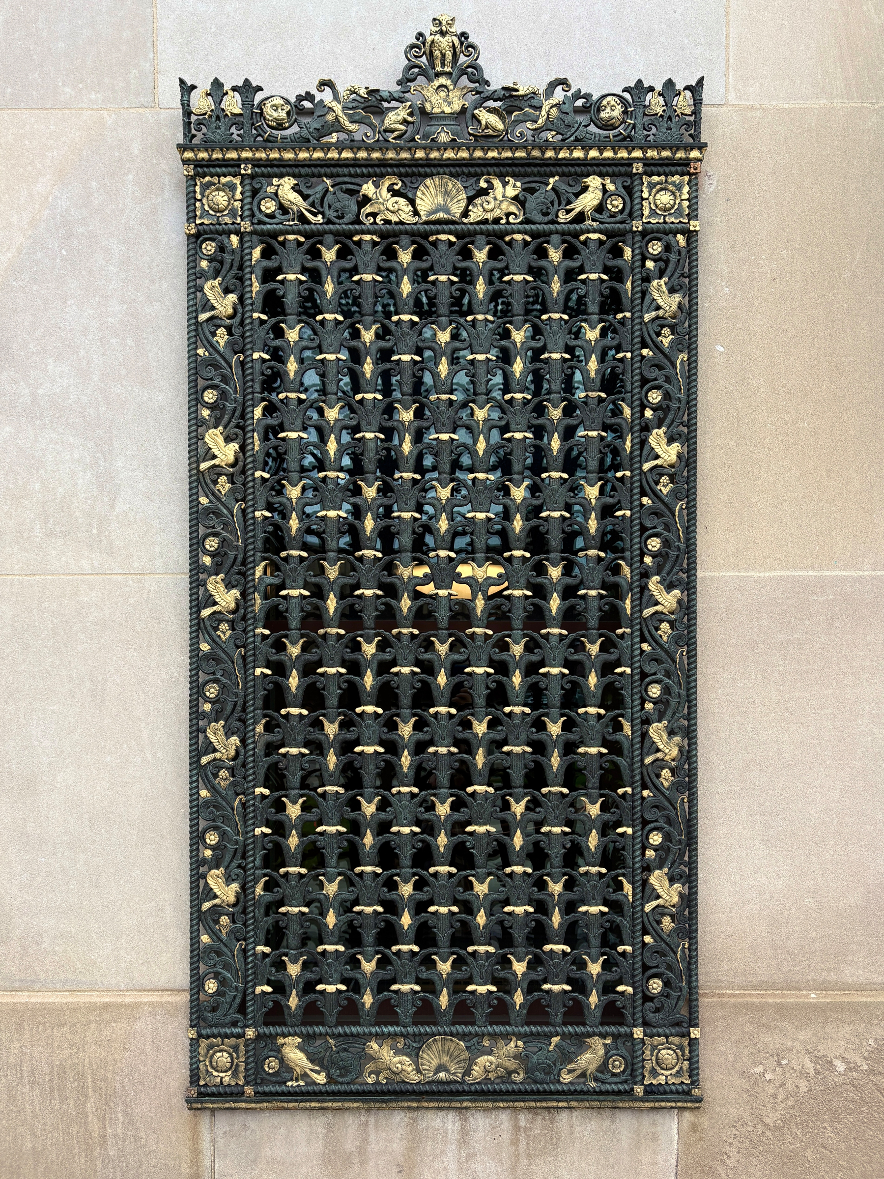 An ornate, rectangular wrought iron grate with intricate gold embellishments is mounted on a stone wall.