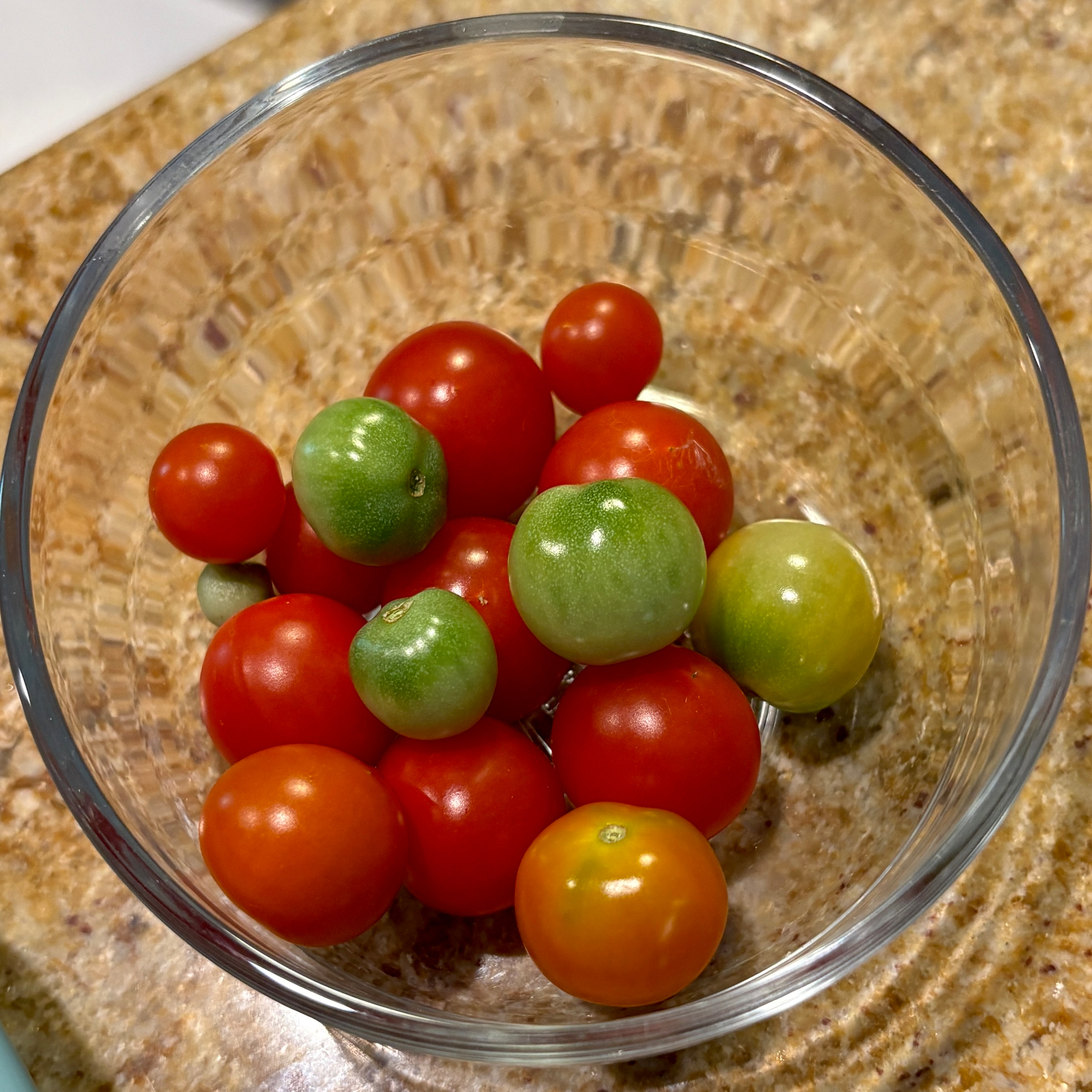 A clear glass bowl contains a mix of ripe red and unripe green cherry tomatoes on a granite countertop.