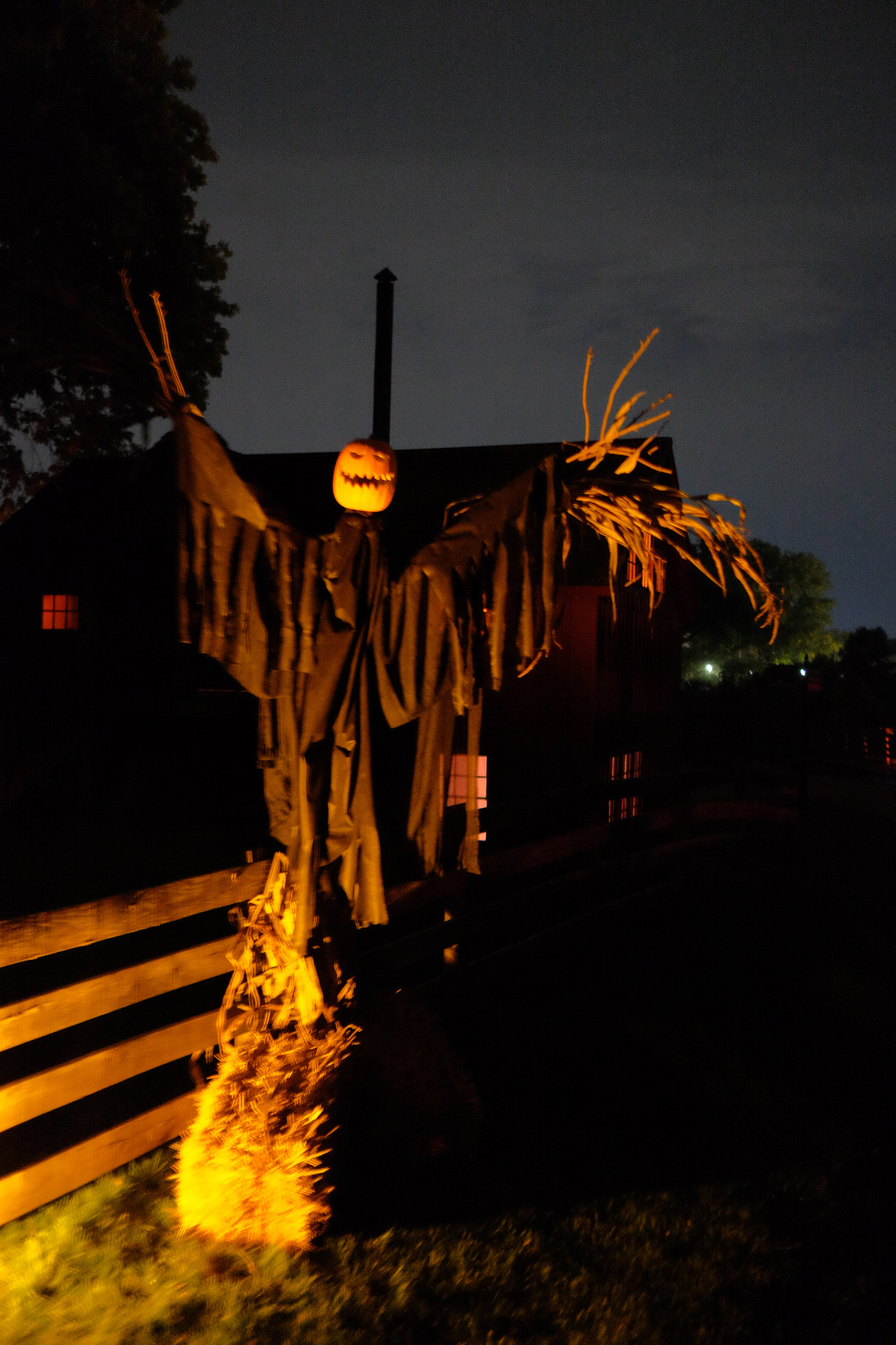 A scarecrow with a pumpkin head, tattered black clothing, and outstretched arms stands illuminated at night near a fence.