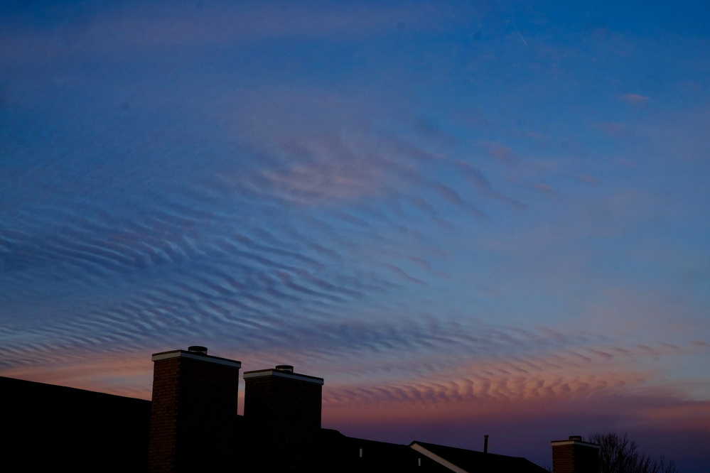 Wispy, wave-like clouds fill the sky at sunset above silhouetted rooftops and chimneys.