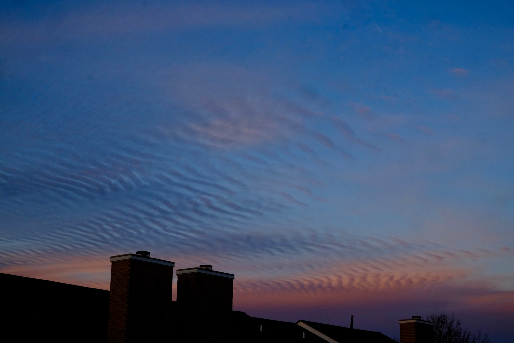 Wispy, wave-like clouds fill the sky at sunset above silhouetted rooftops and chimneys.