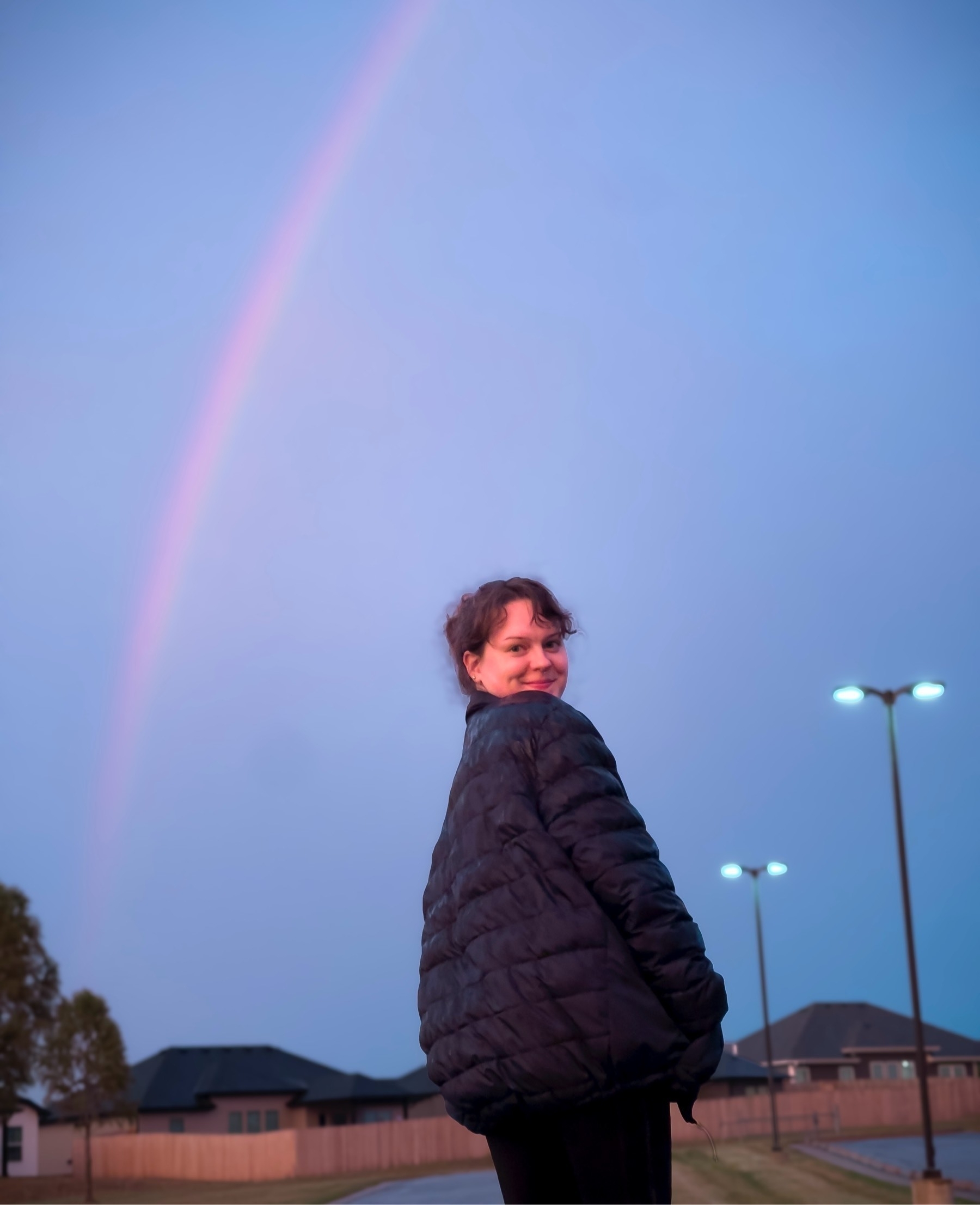 Young woman turned back to look at camera beneath a dusk rainbow.
