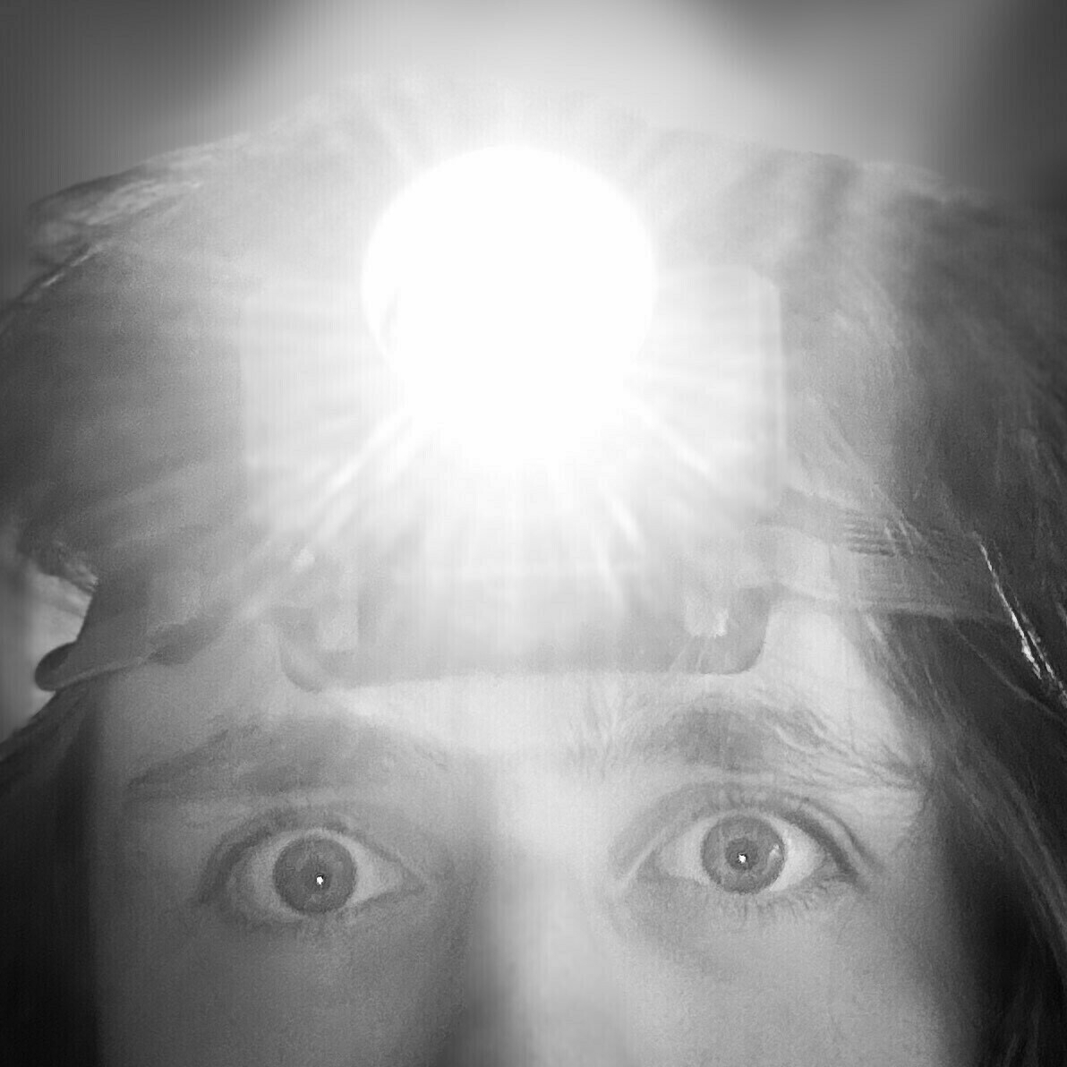 Close-up of a person's eyes with a bright light on their forehead, possibly from a headlamp, in black and white.