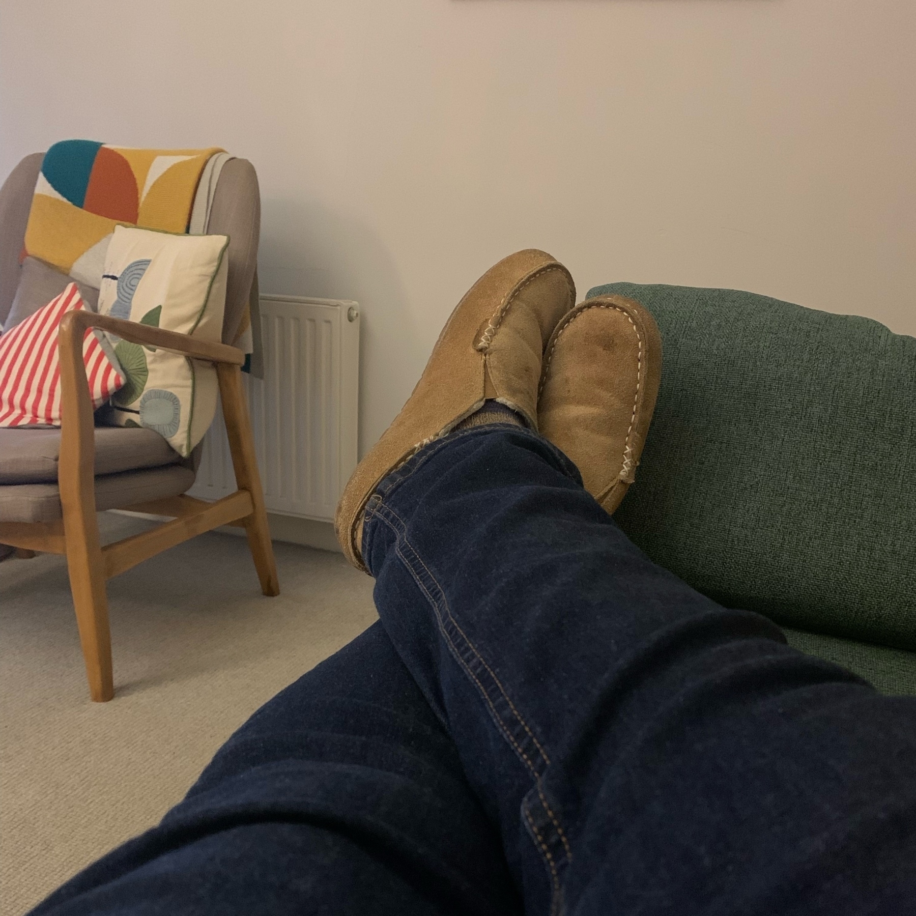 Crossed legs in dark blue jeans and suede slippers. Armchair in background