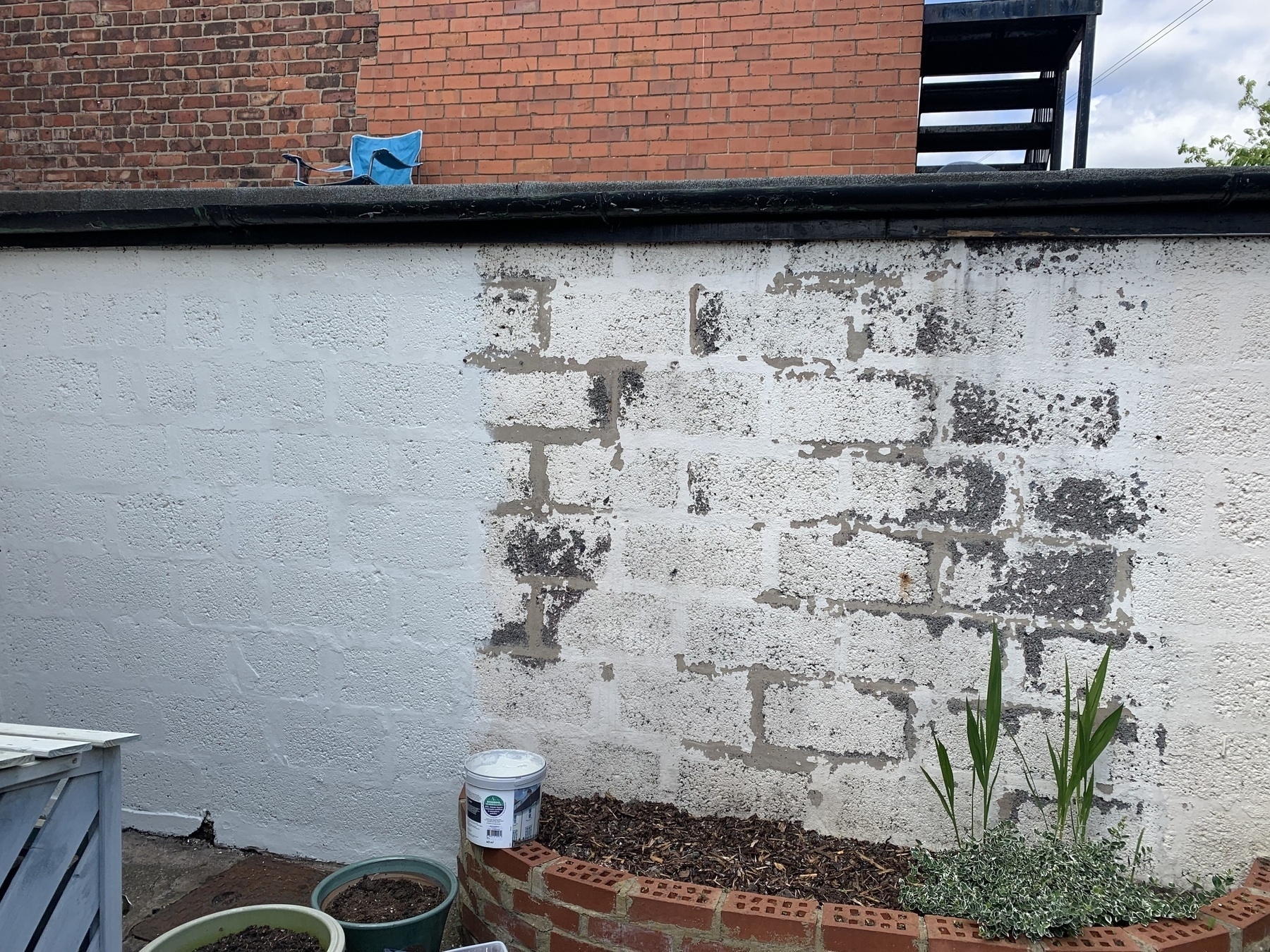 External breeze block wall with flaking paint and bare patches in the centre. Painting in progress from left to right with clear line marking done and not done. Open paint can in view on a low brick wall.