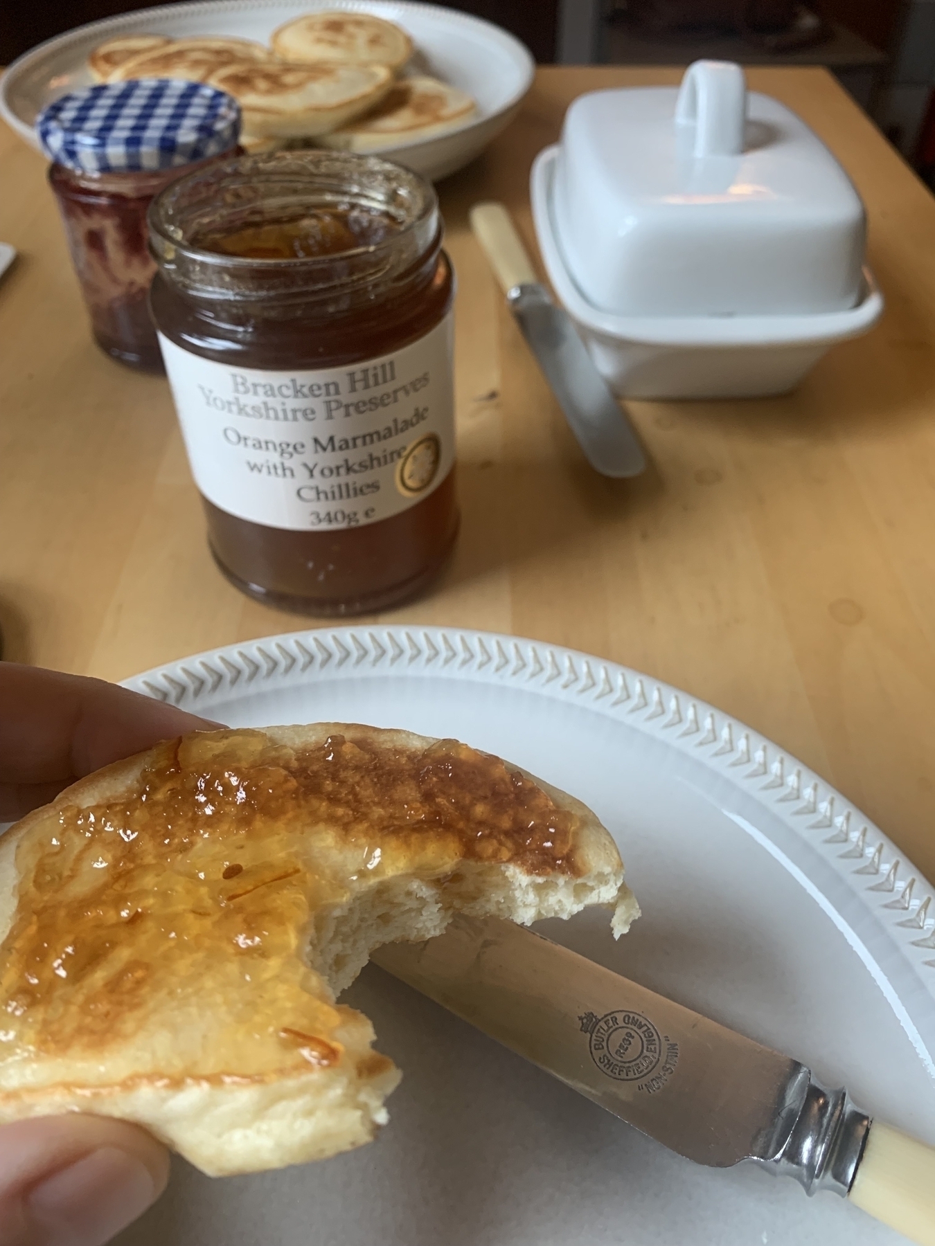 Drop scone with a bite take from it covered in a thin layer of preserve. Open jar of orange marmalade with Yorkshire chillies in the background.
