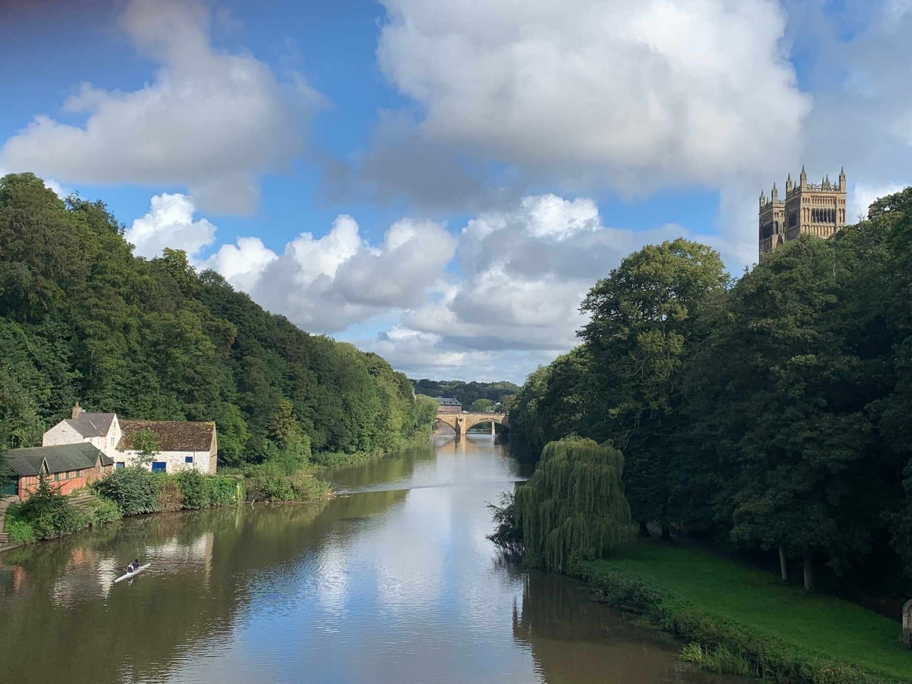 A view from a bridge crossing the River Wear. Trees line both banks. A double scull boat is in the water. Durham Cathedral peaks out from behind the treetops on the right riverbank.