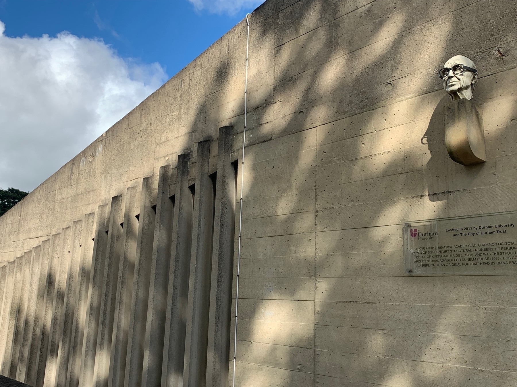 Side of a concrete building with multiple narrow vertical windows and a small bust of the architect Ove Arup sitting on a plinth attached to the wall.