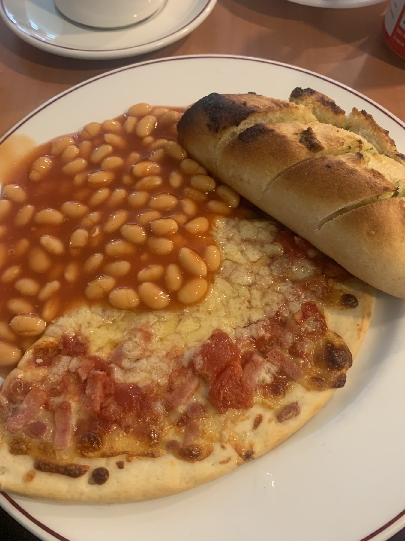 White china plate filled with a huge slice of pizza, half a garlic bread baguette and a mound of baked beans.