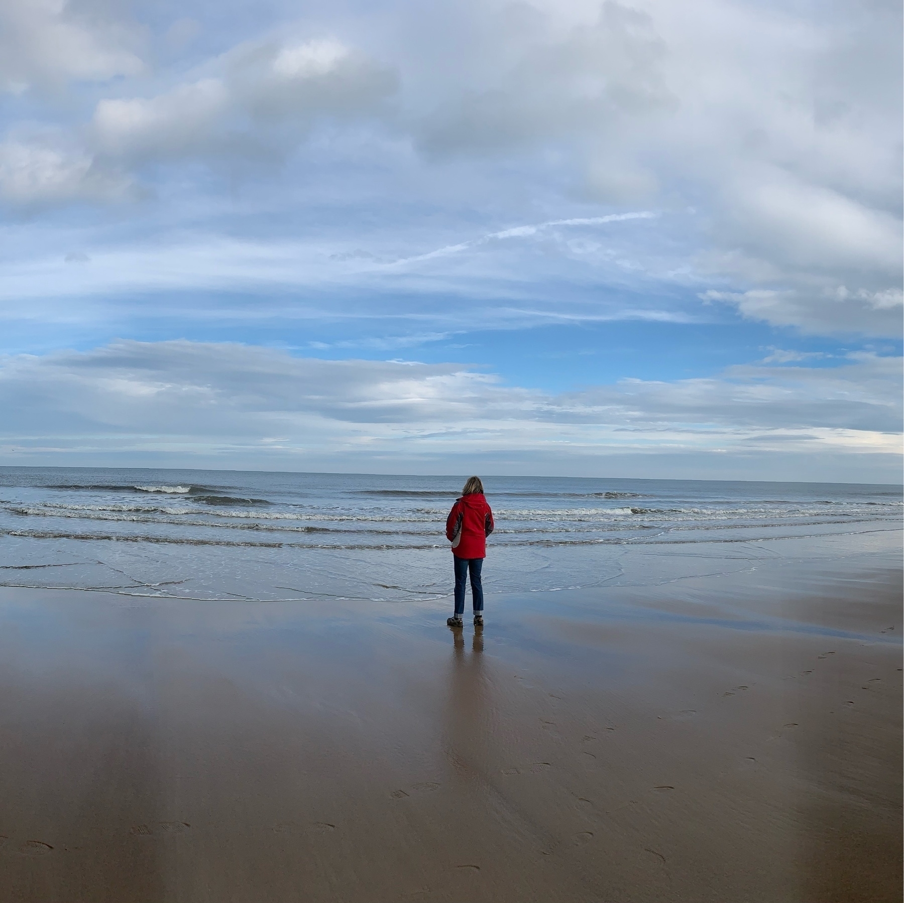 Woman in a red coat stands on the sand looking out to sea