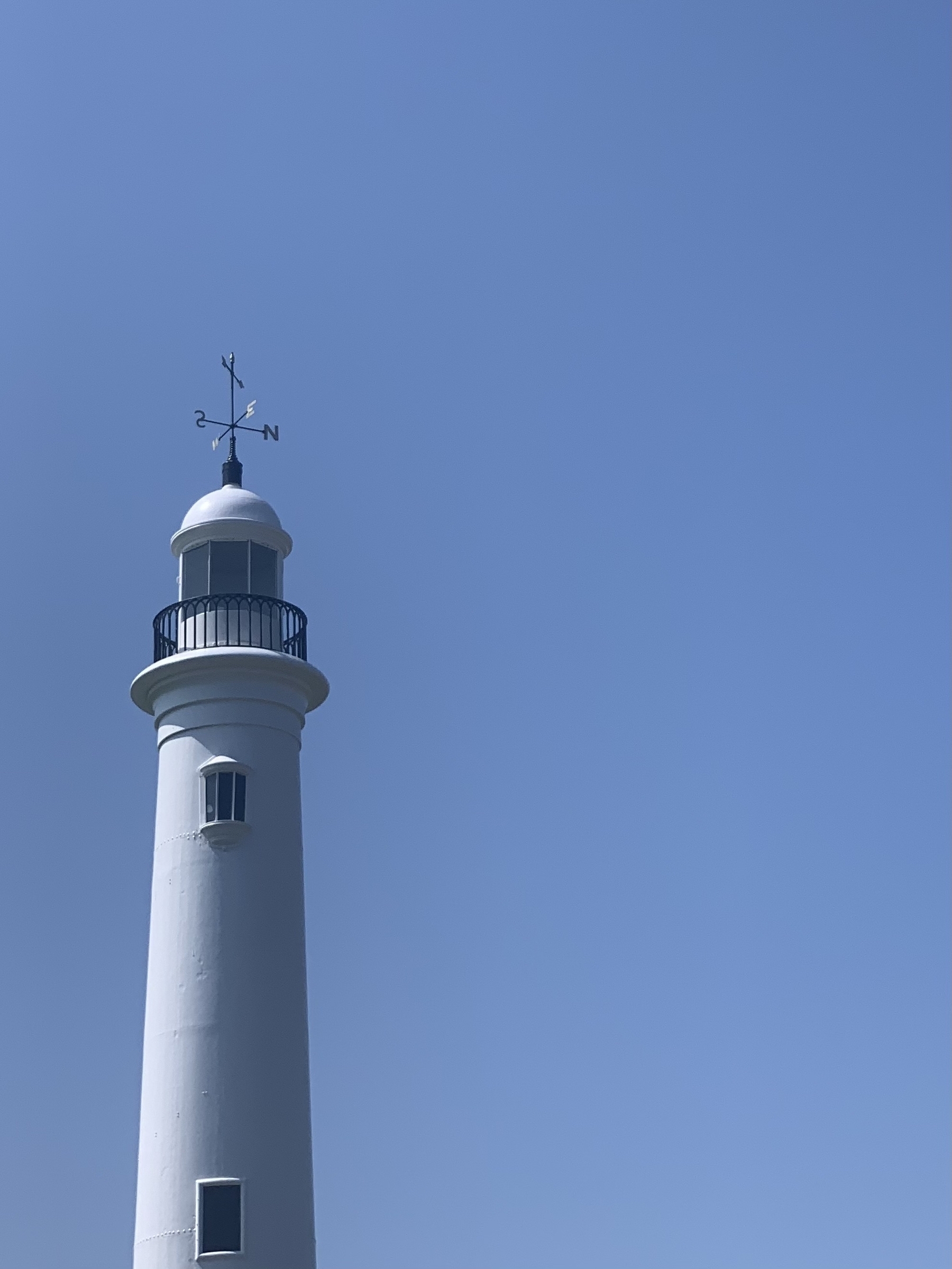 White lighthouse tower with weather vane on it’s roof against a bright blue sky.