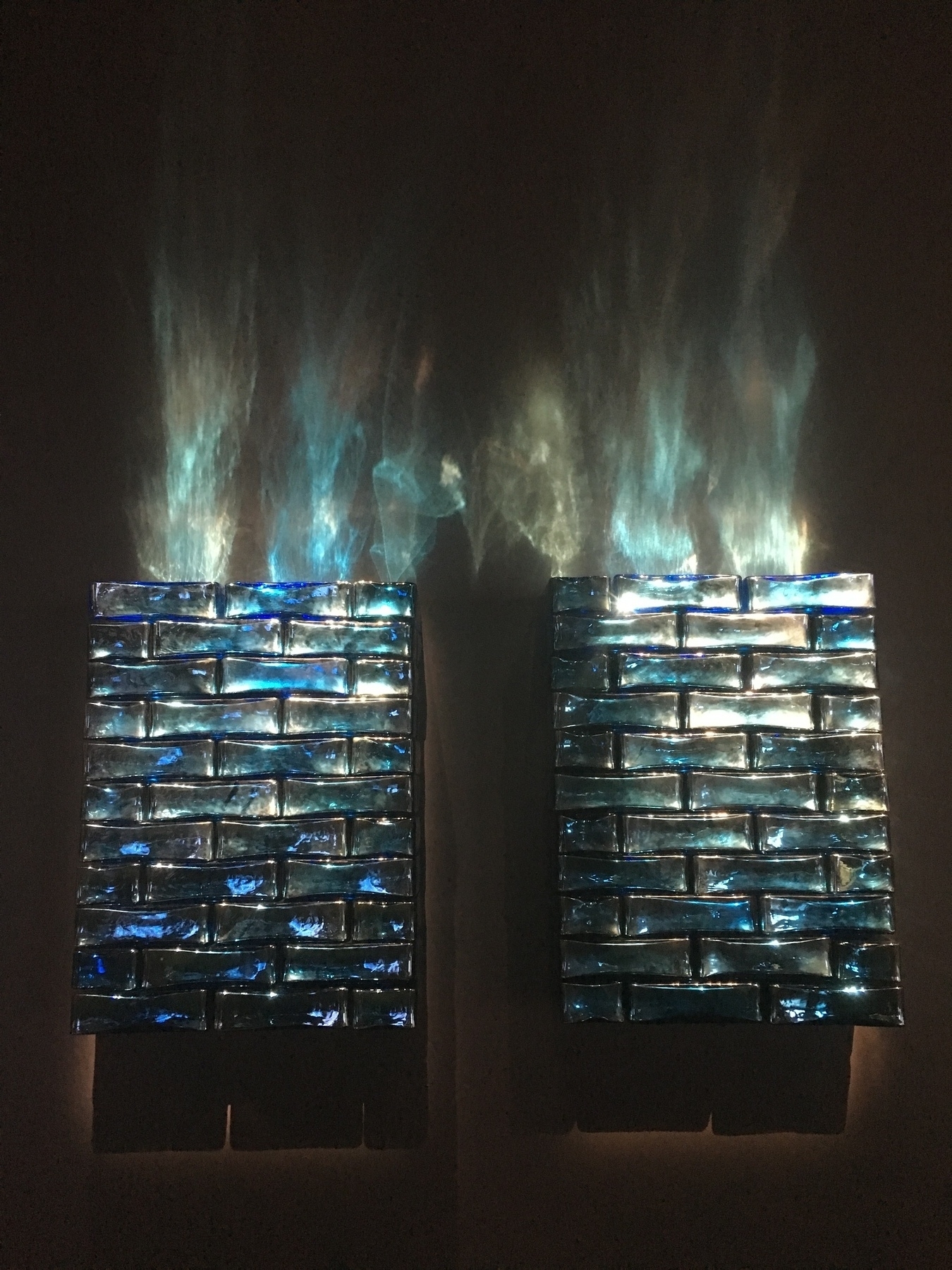 Two wall mounted rectangular objects made of glass blocks in various shades of blue with dispersed lights projected upwards from the top of them.