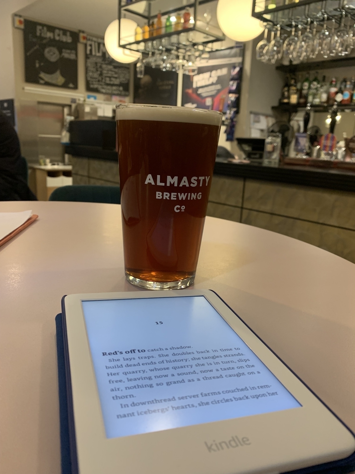Pint of brown ale in an Almasty Brewing Co glass sitting on a white table behind an open Kindle.