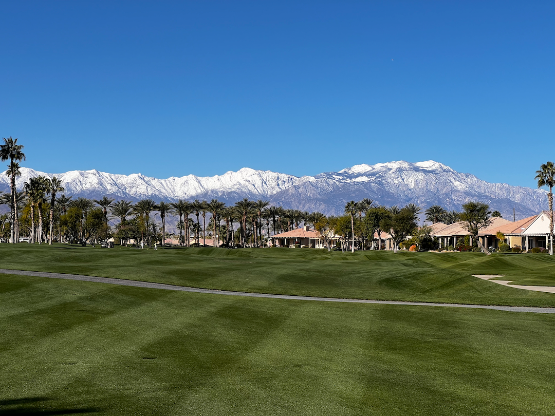 A lush green golf course is set against a backdrop of snow-capped mountains and clear blue sky.