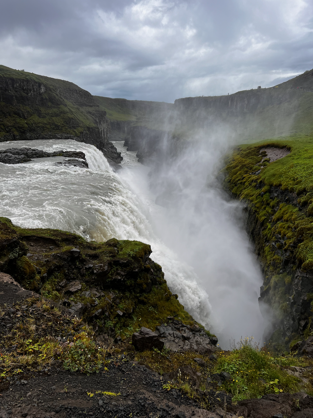 A powerful waterfall cascades into a rocky gorge surrounded by lush green cliffs under a cloudy sky.